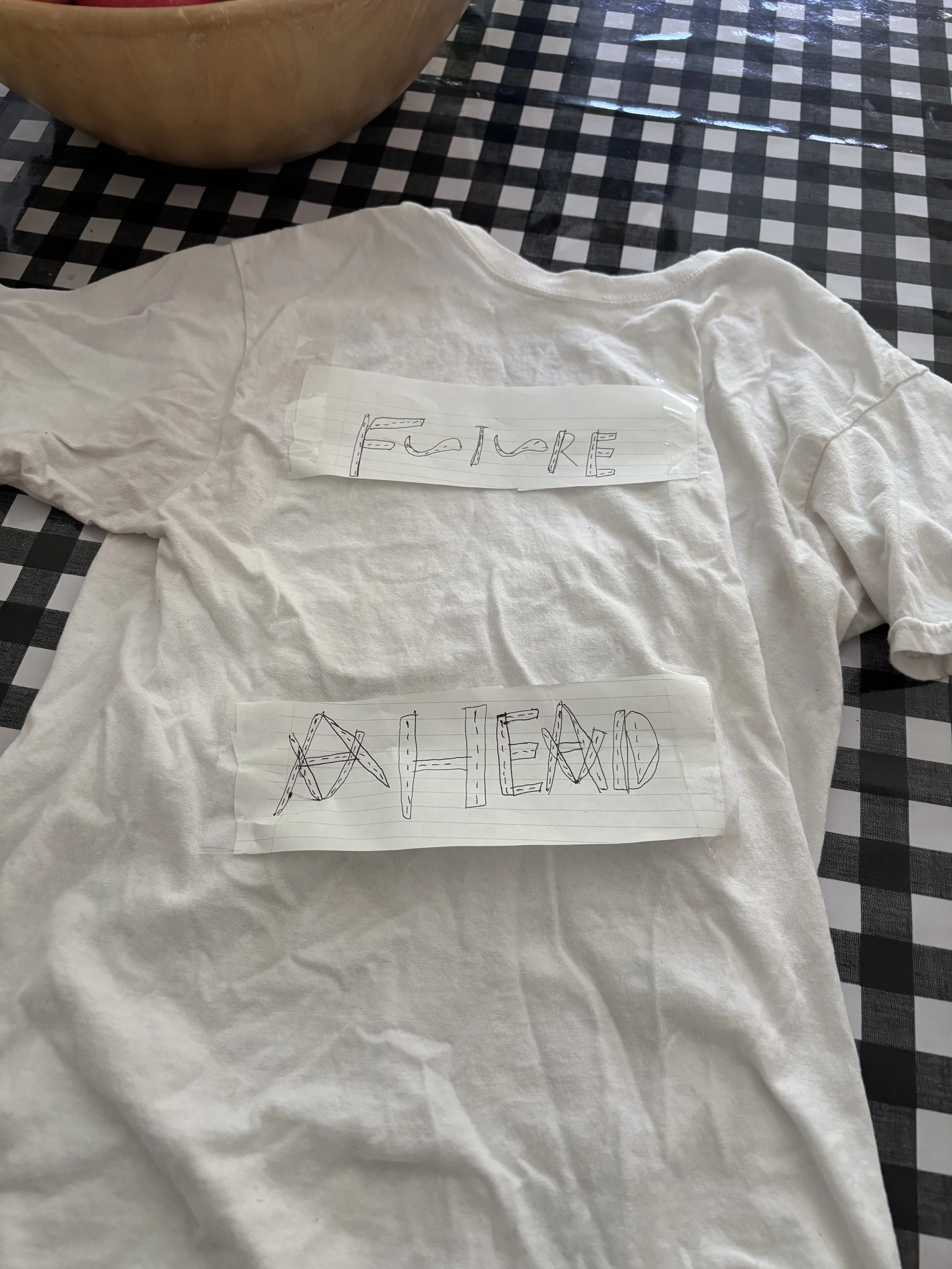 A white T-shirt on a checkered table, with two sheets of lined paper taped to it. One sheet has the word 'FUTURE' written in stylized letters, and the other has the word 'BEYOND' written in large, block letters.