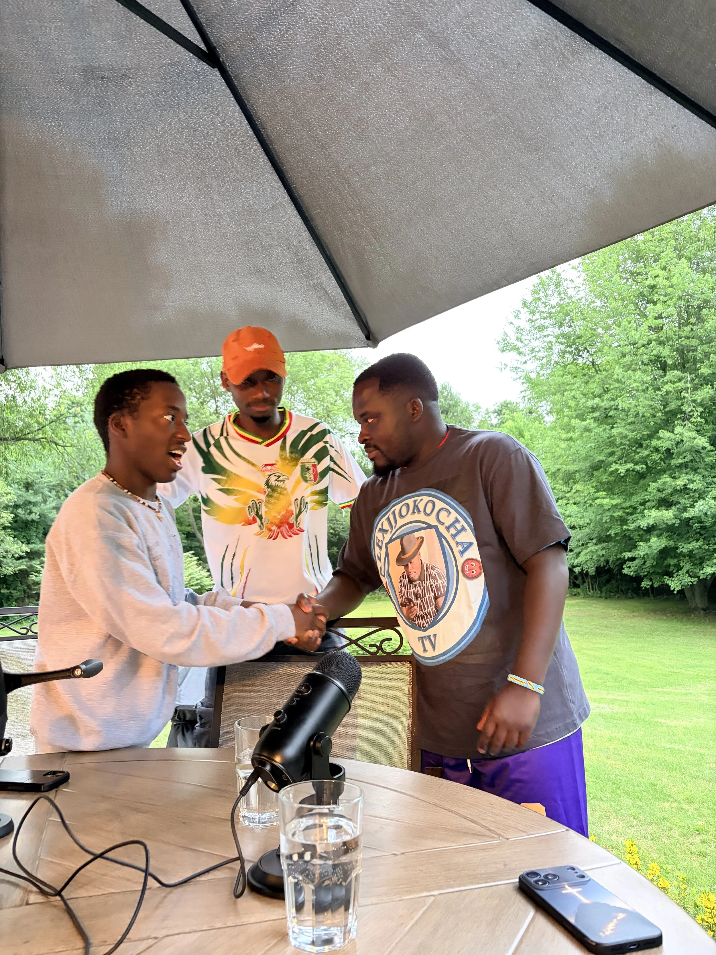 Three men are outside under a large umbrella. Two are shaking hands, one looks on. The man on the left is wearing a light gray hoodie, the man in the center is wearing a colorful shirt with a dragon and an orange cap, and the man on the right is wearing a gray T-shirt with a graphic and purple shorts. There are glasses of water, a microphone, a phone, and other objects on a table in the foreground. Green trees and grass are in the background.
