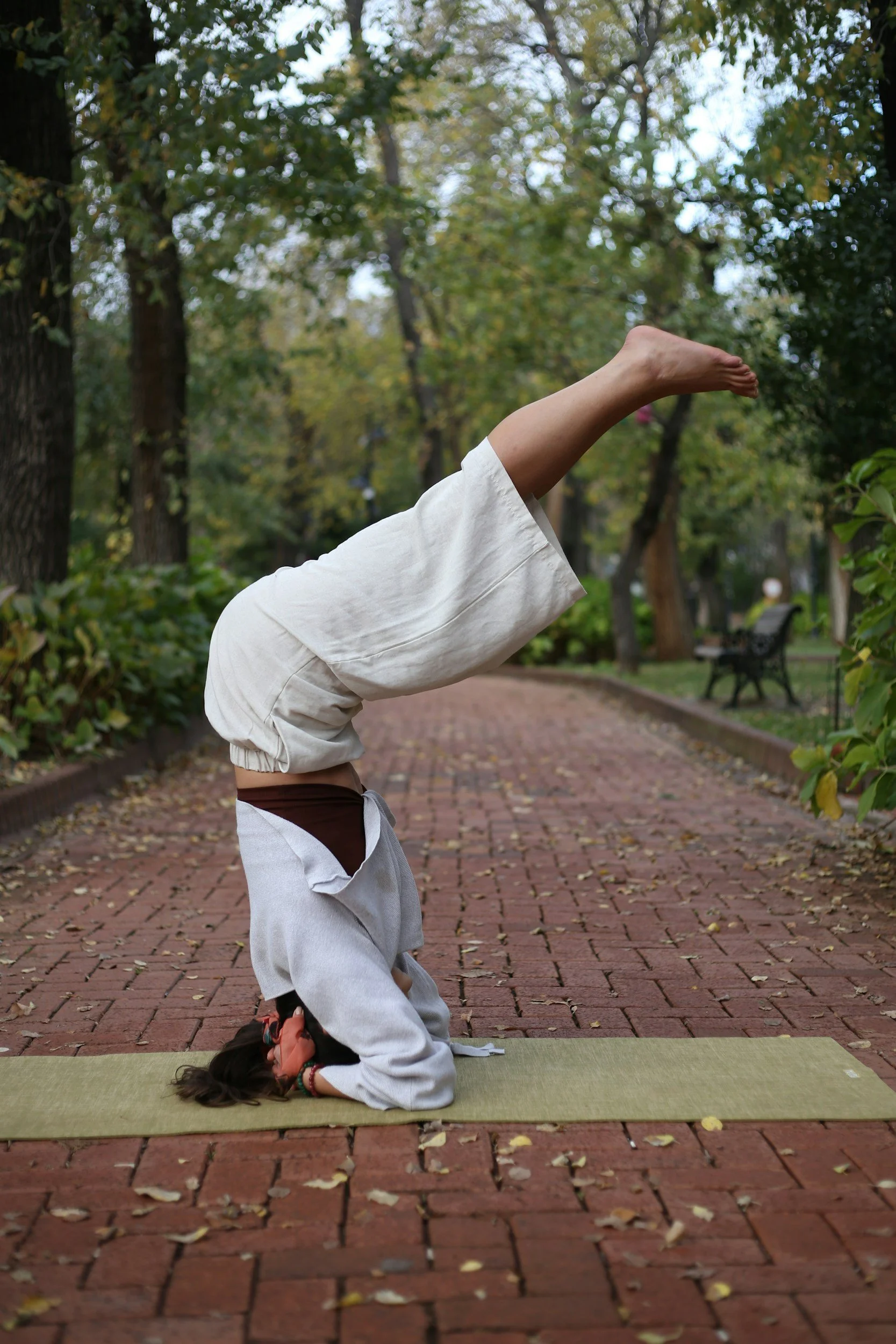 A person practicing yoga outdoors on a green mat, performing a headstand with another person in a child's pose beneath them, surrounded by trees and fallen leaves on a brick pathway.