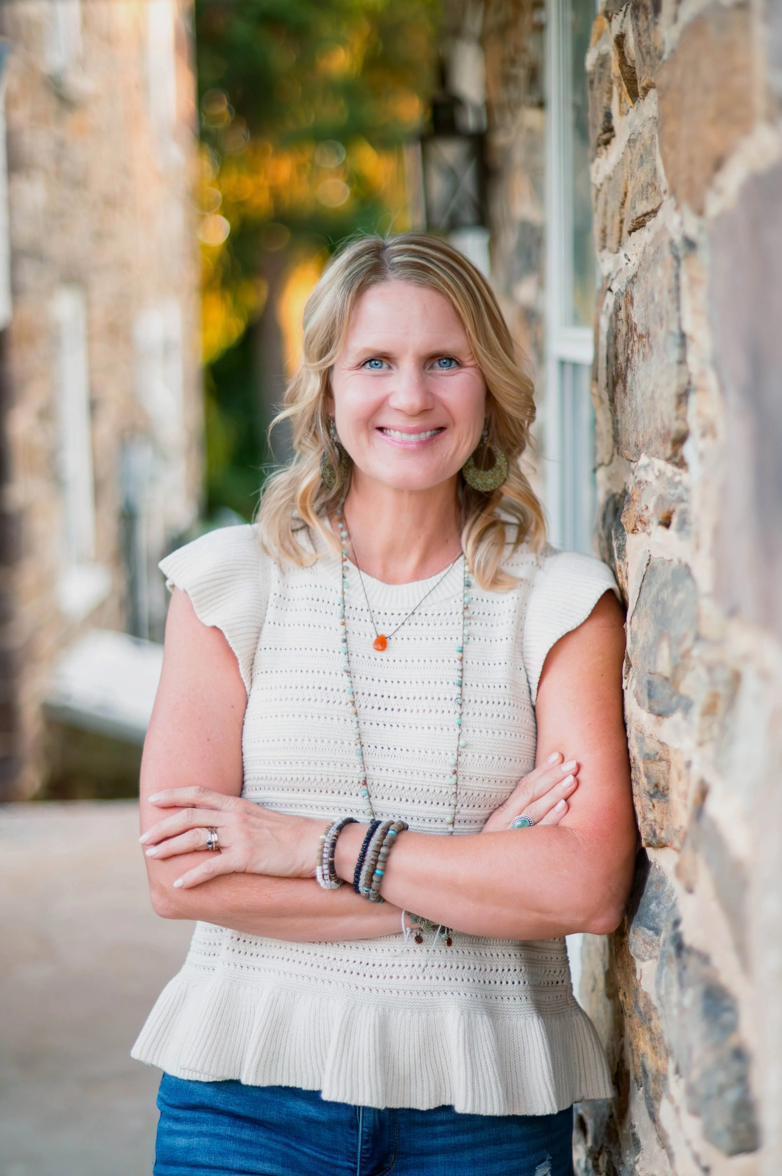 Portrait of a woman with blonde hair and blue eyes standing outdoors near a stone wall, smiling with arms crossed, wearing a white sleeveless knit top and layered jewelry.