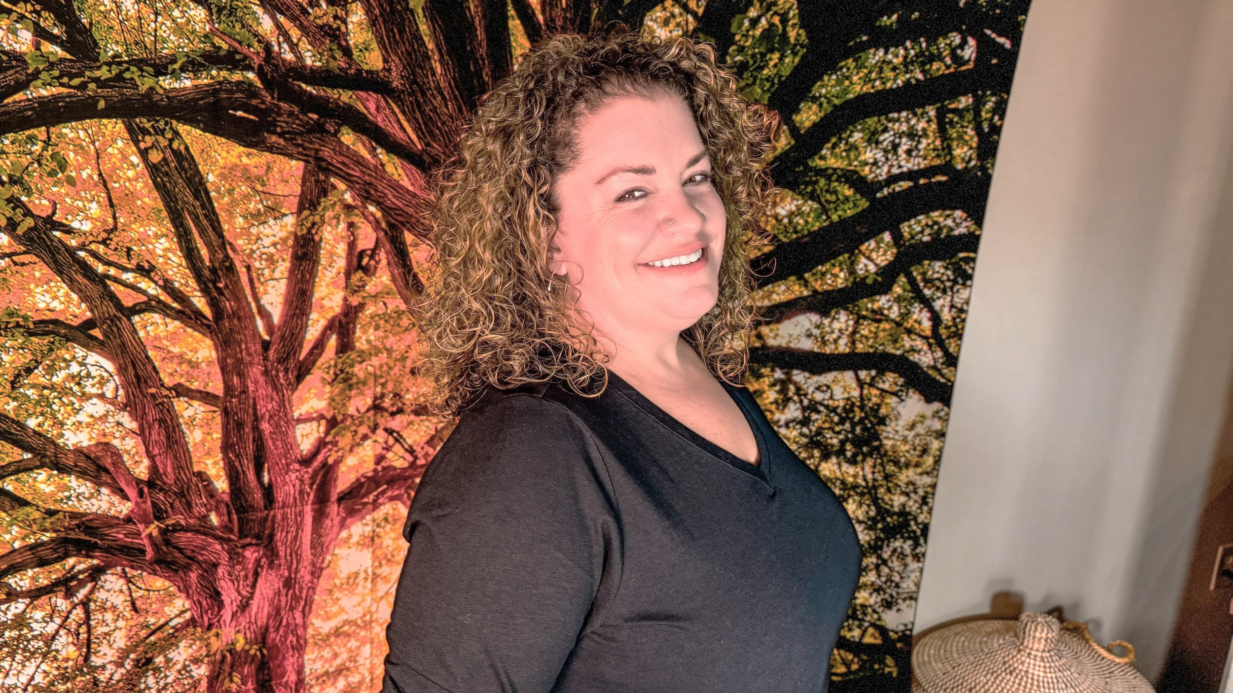 A woman with curly hair, smiling, wearing a black top, standing in front of a colorful autumn tree backdrop.