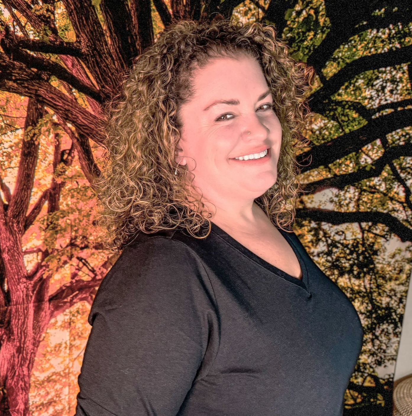 A woman with curly hair smiling in front of a backdrop of a large tree with autumn leaves.