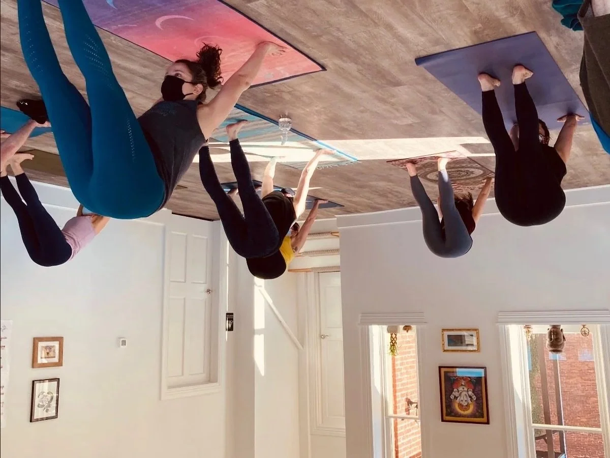 Three women participating in a yoga class, practicing poses on yoga mats in a bright, well-lit room with windows and artwork on the walls.
