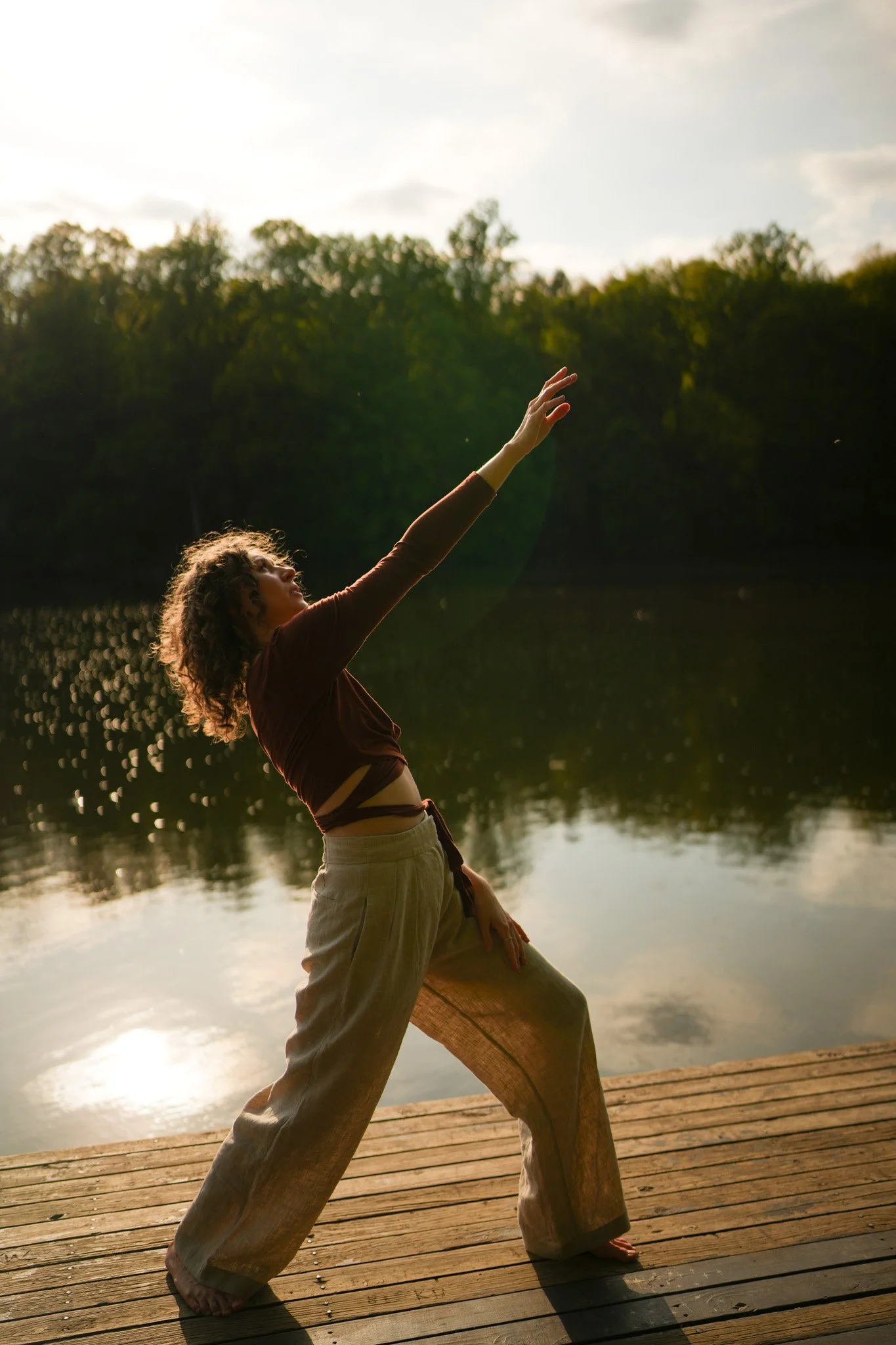 A person with curly hair practicing yoga or stretching on a wooden dock beside a calm body of water, with trees in the background and the sky partly cloudy.
