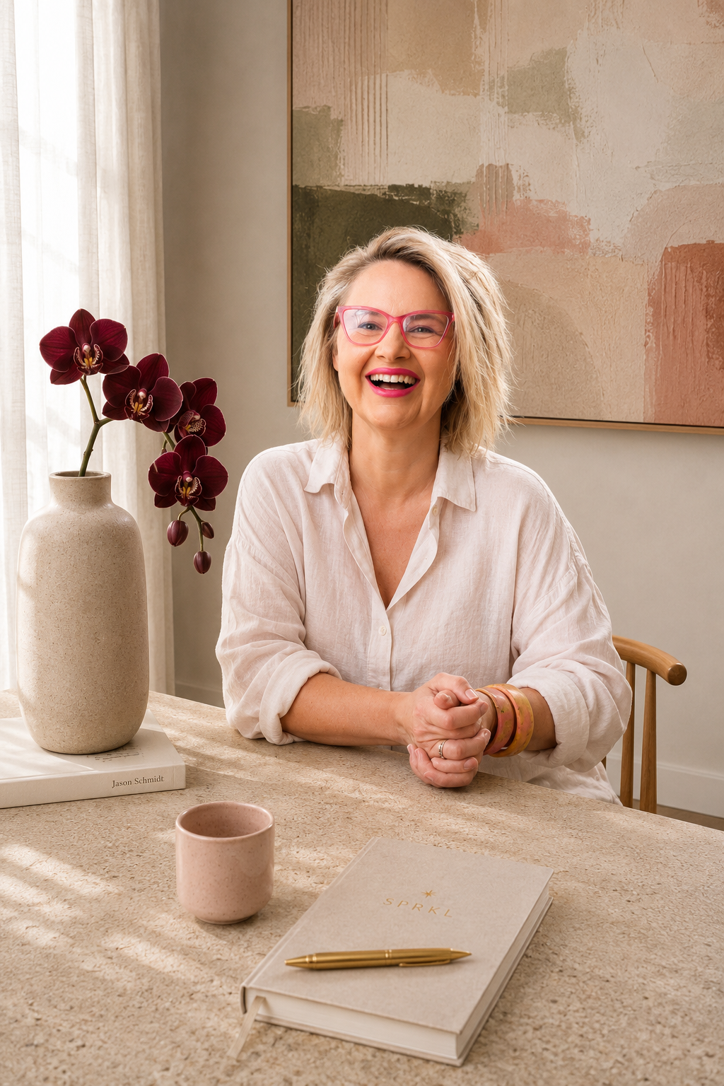 Rachael Karlyl wearing pink glasses, a light beige shirt, and bracelets, sitting at a beige textured table next to a white vase with burgundy orchids, a pink mug, and a cream-colored planner with 'SPRKL' on the cover. Leader. Coach. Training. Exec.