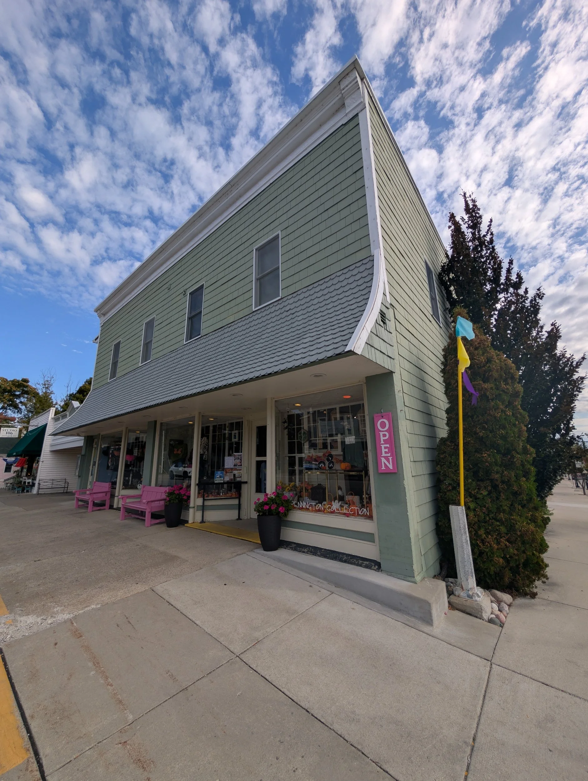 A two-story light green building with large windows and a storefront on the ground level. Pink benches and potted flowers are outside. An 'Open' sign hangs on the window, and colorful flags are on a pole nearby. The sky is partly cloudy.