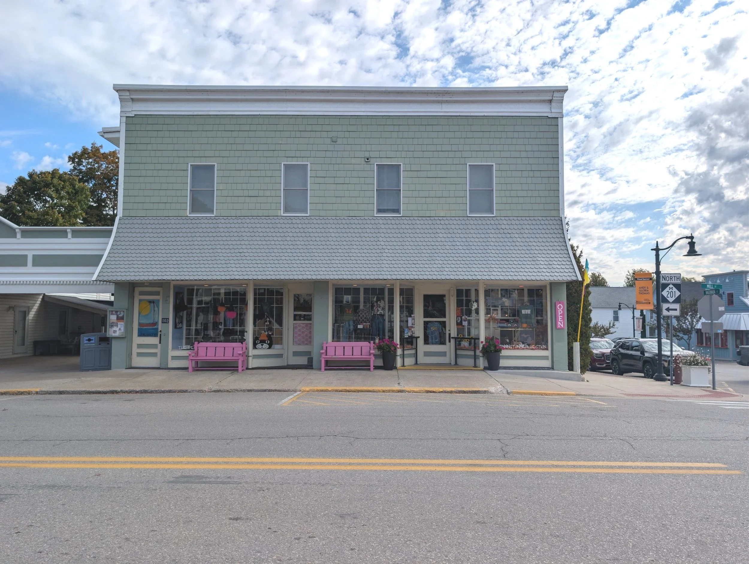 Front view of a two-story storefront building with light green shingle exterior and large windows, pink benches outside, and cloudy sky overhead.