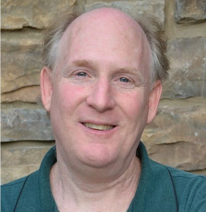 A close-up of a middle-aged man with light skin, light-colored hair, and a friendly expression, standing in front of a stone wall.