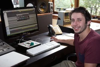 Young man sitting at a music production workstation with a computer, mixing console, speakers, and recording equipment in a well-lit room.