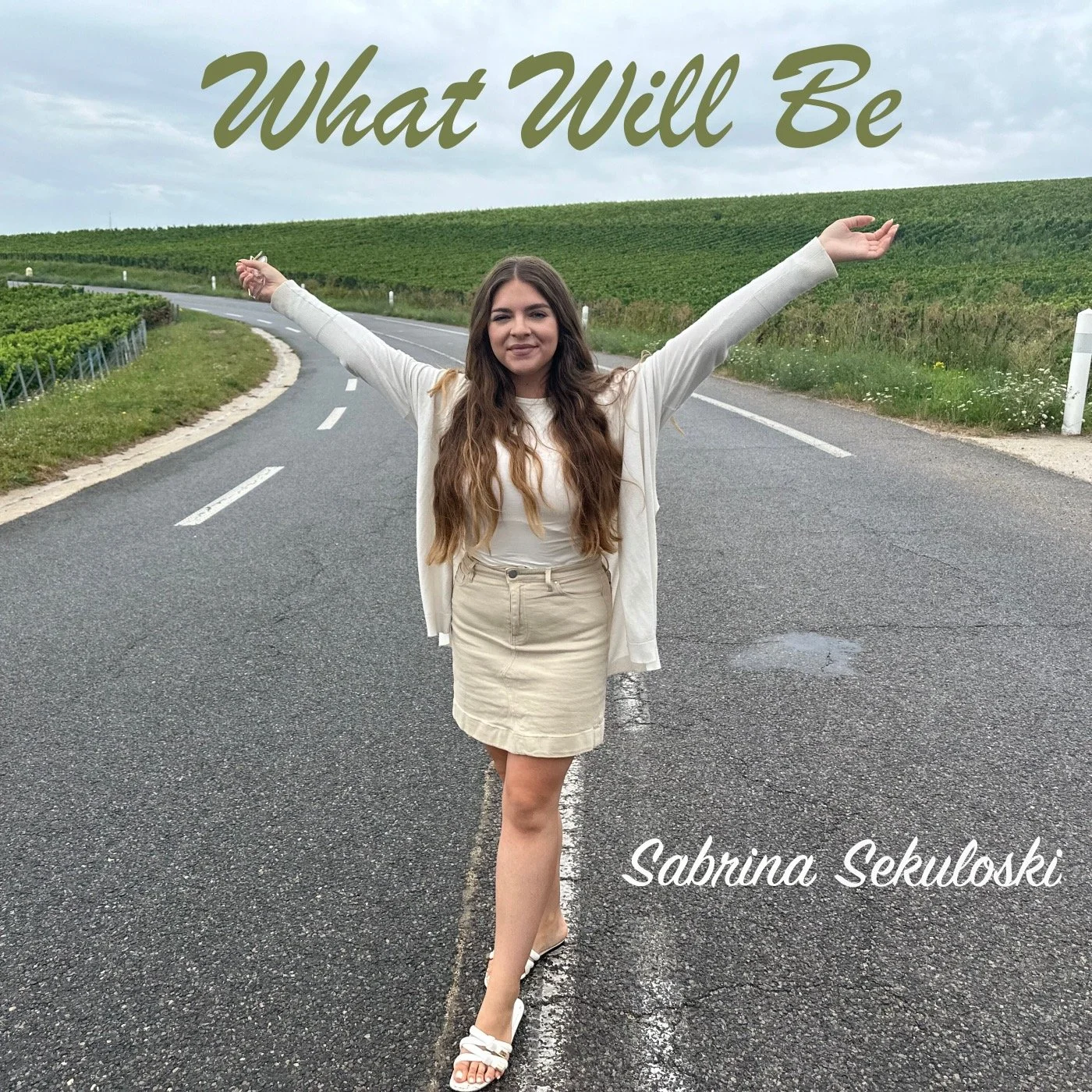A woman standing in the middle of a rural road with arms outstretched. Text overlay says, "What Will Be," and at the bottom, it says, "Sabrina Sekulowski."