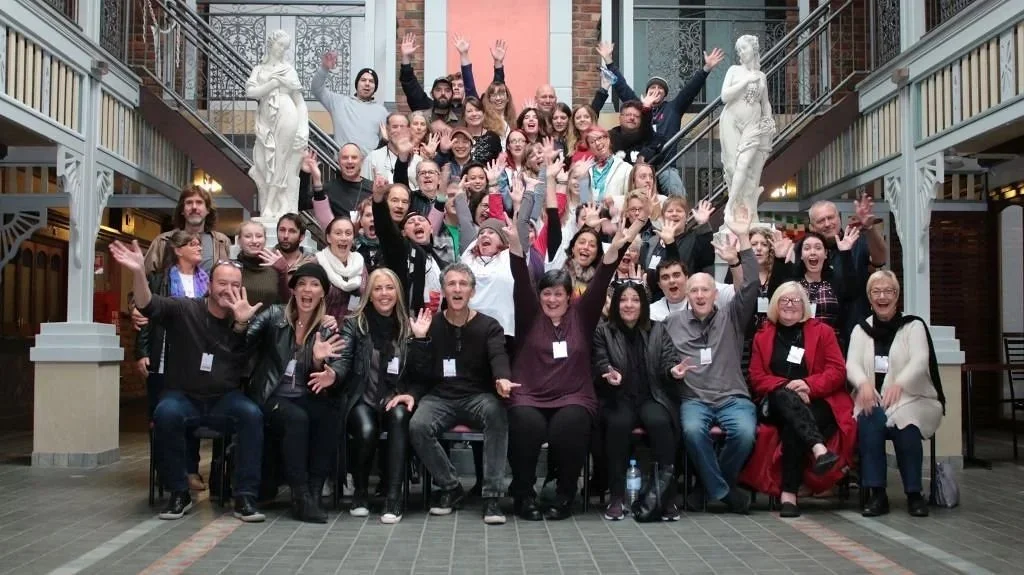 Group of diverse people at the Australian Songwriters Conference cheering and smiling in a spacious, decorated venue with statues and balcony railings.
