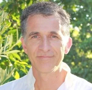 A middle-aged man with short, curly gray hair outdoors, smiling softly with greenery and sunlight in the background.