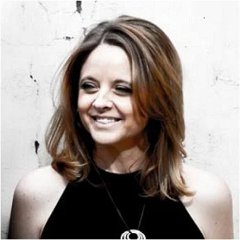 A smiling woman with shoulder-length brown hair, wearing a black sleeveless top and a necklace, standing against a white textured wall.
