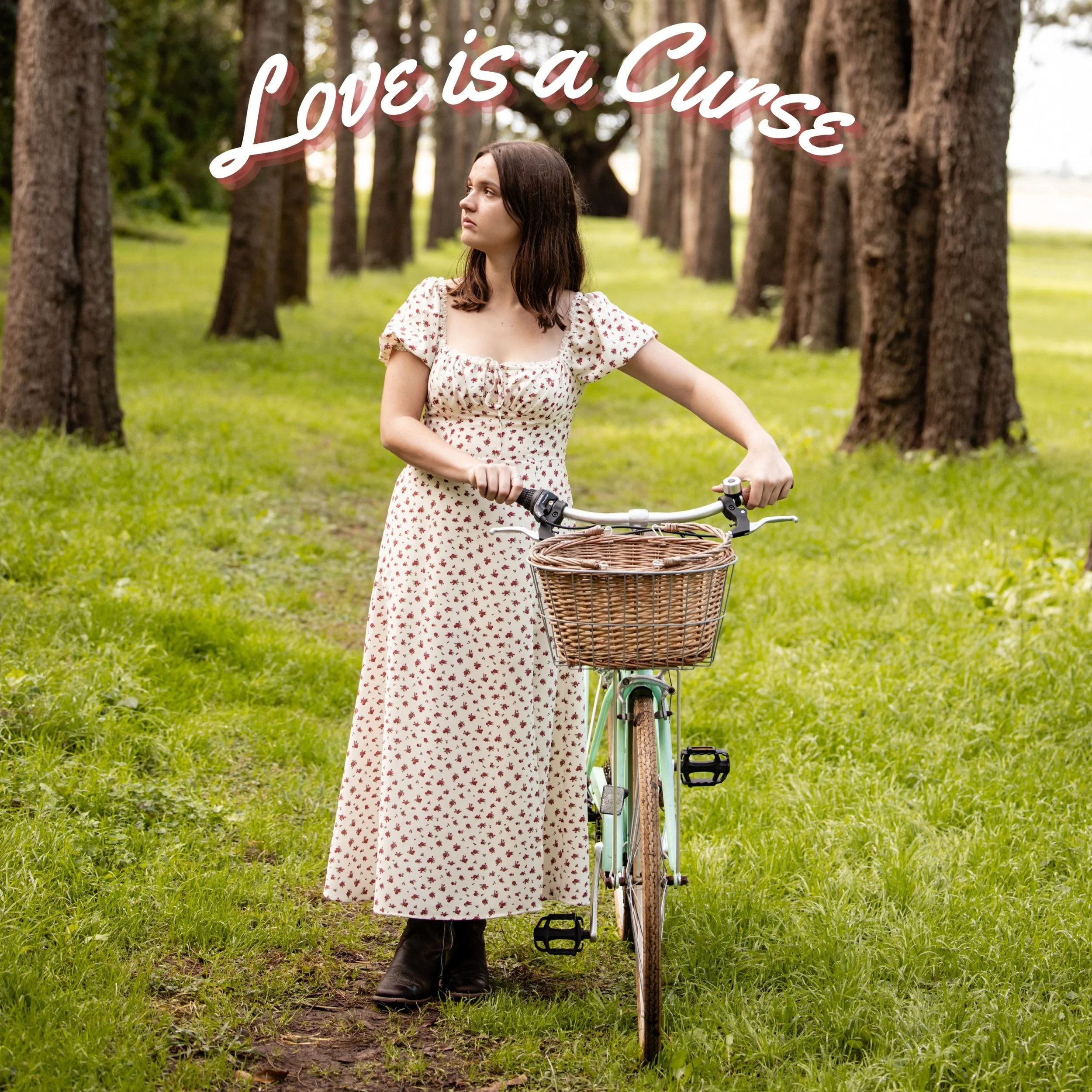 A woman standing in a wooded area holding a bicycle with a wicker basket, wearing a white dress with red floral pattern, and looking to her left. There is text overlay that reads 'Love is a Curse' in white and red script font.