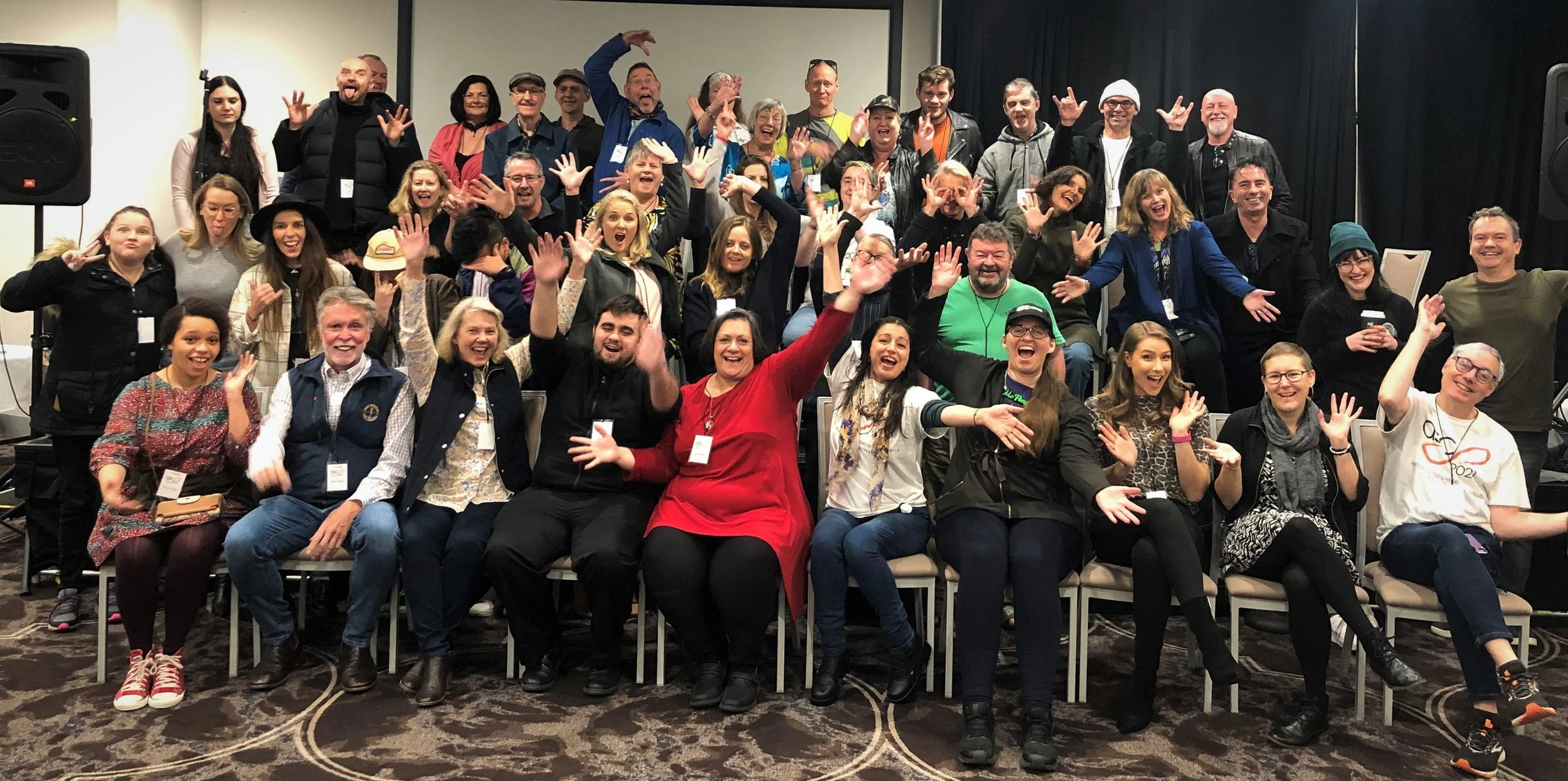 A large group of people gathered together indoors for a group photo, all smiling and waving.