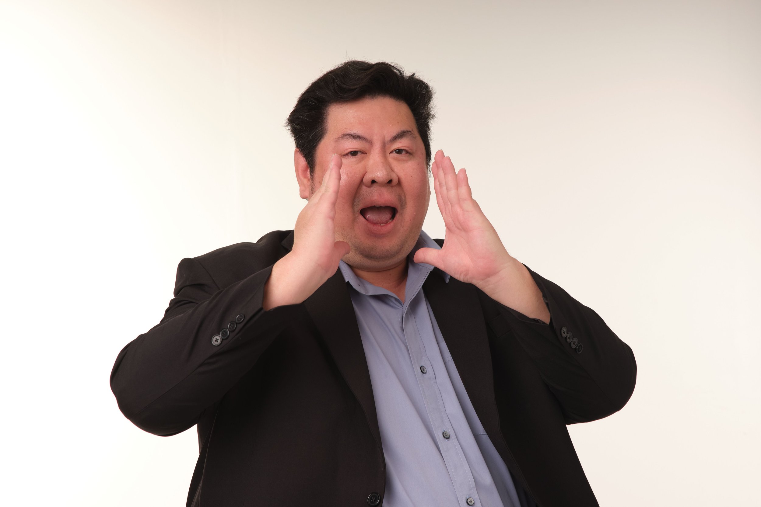 A man in a suit making a loud gesture with hands near his face, mouth open, standing against a plain white background.