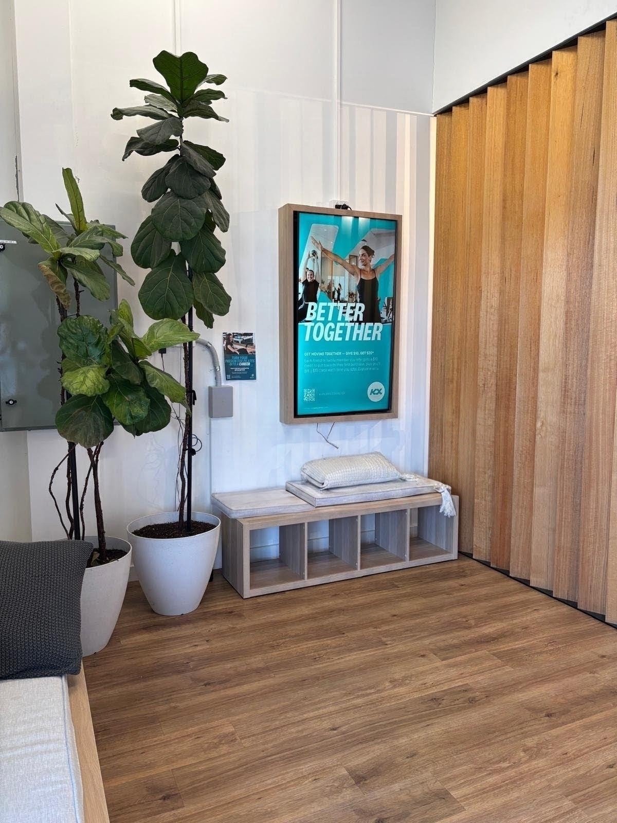 Indoor waiting area with large potted plants, a digital advertisement display with the text 'Better Together', a wooden bench with a folded towel, and wooden wall paneling.