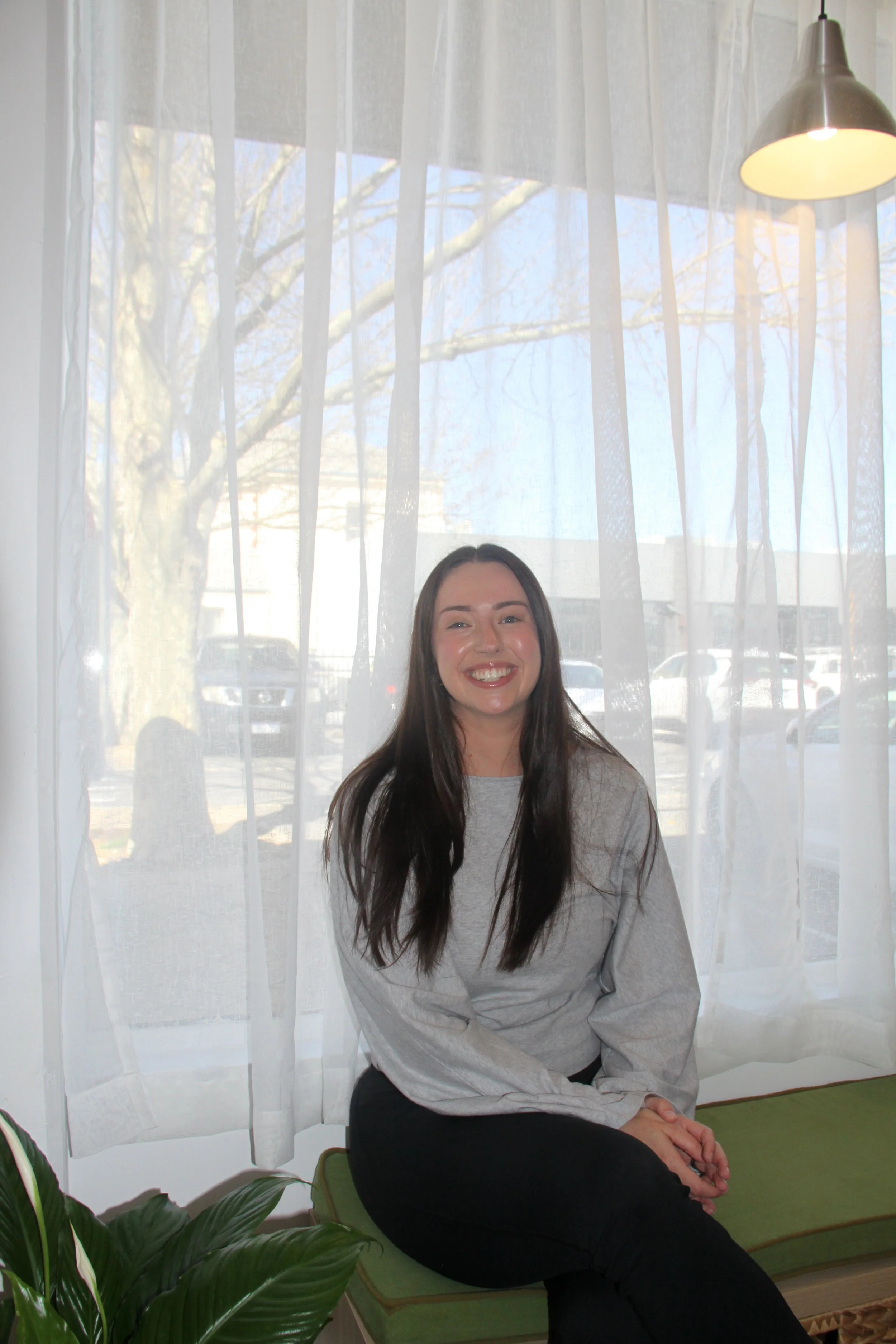 A woman with long dark hair smiling, sitting on a green bench in front of a window with white sheer curtains. Outside the window, leafless trees and parked cars are visible. A silver hanging lamp is above her right shoulder, and a potted plant with broad green leaves is at the bottom left corner of the image.