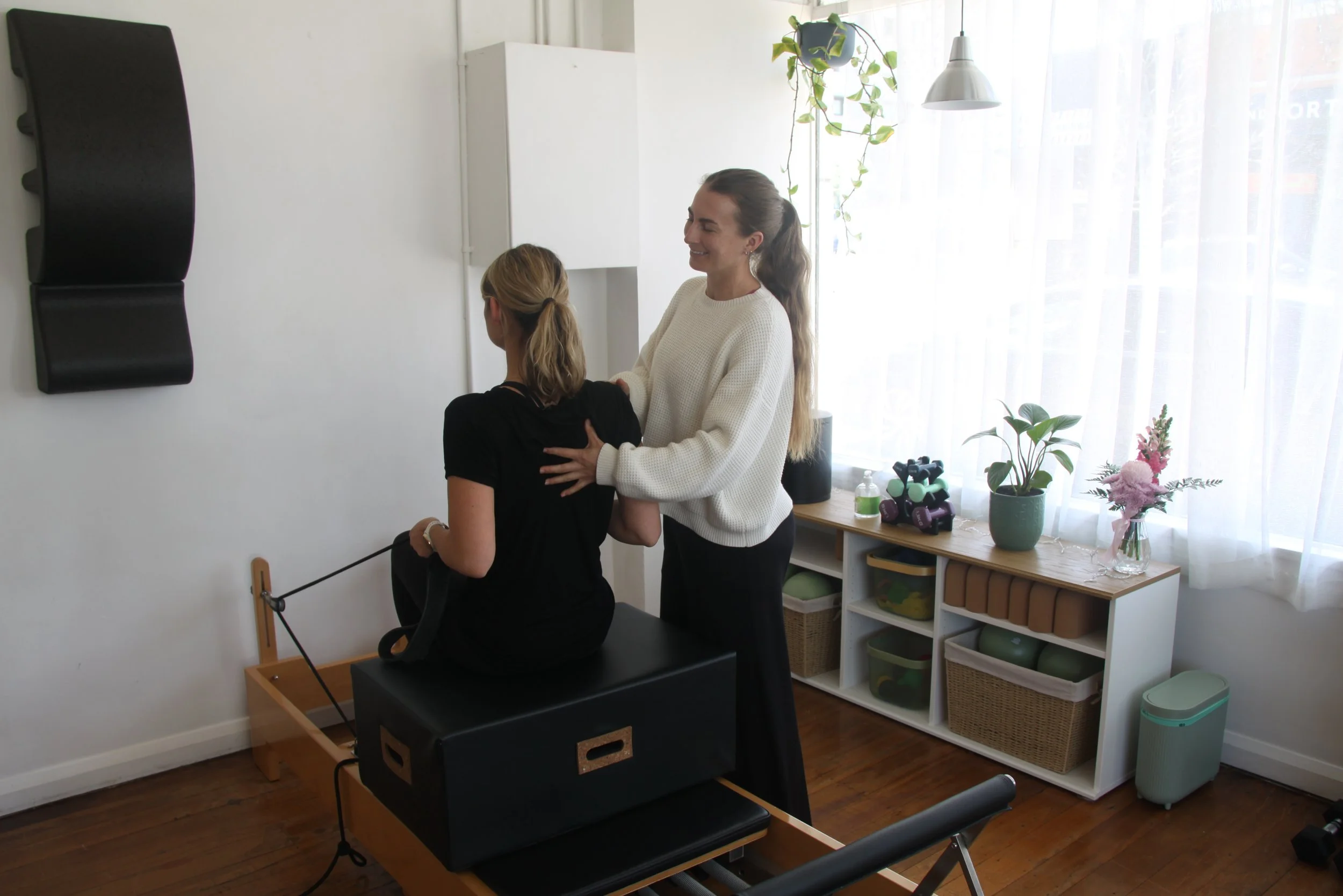 Two women are in a bright room with large windows, one sitting on a Pilates reformer machine performing a fitness exercise and the other standing beside her, smiling and offering support. The room has plants, books, and workout equipment.
