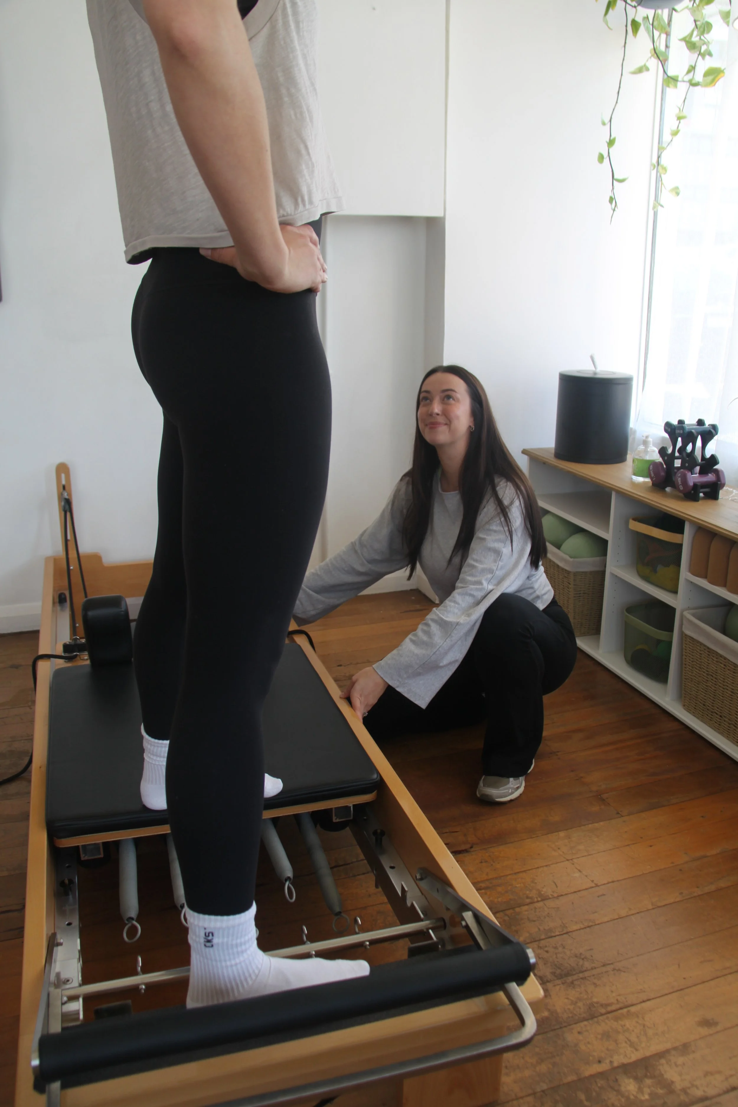 A person standing on a reformer Pilates machine while another person kneels beside them, adjusting or assisting. The room has natural light, wood flooring, and some home gym equipment on a shelf, with hanging plants visible near the window.