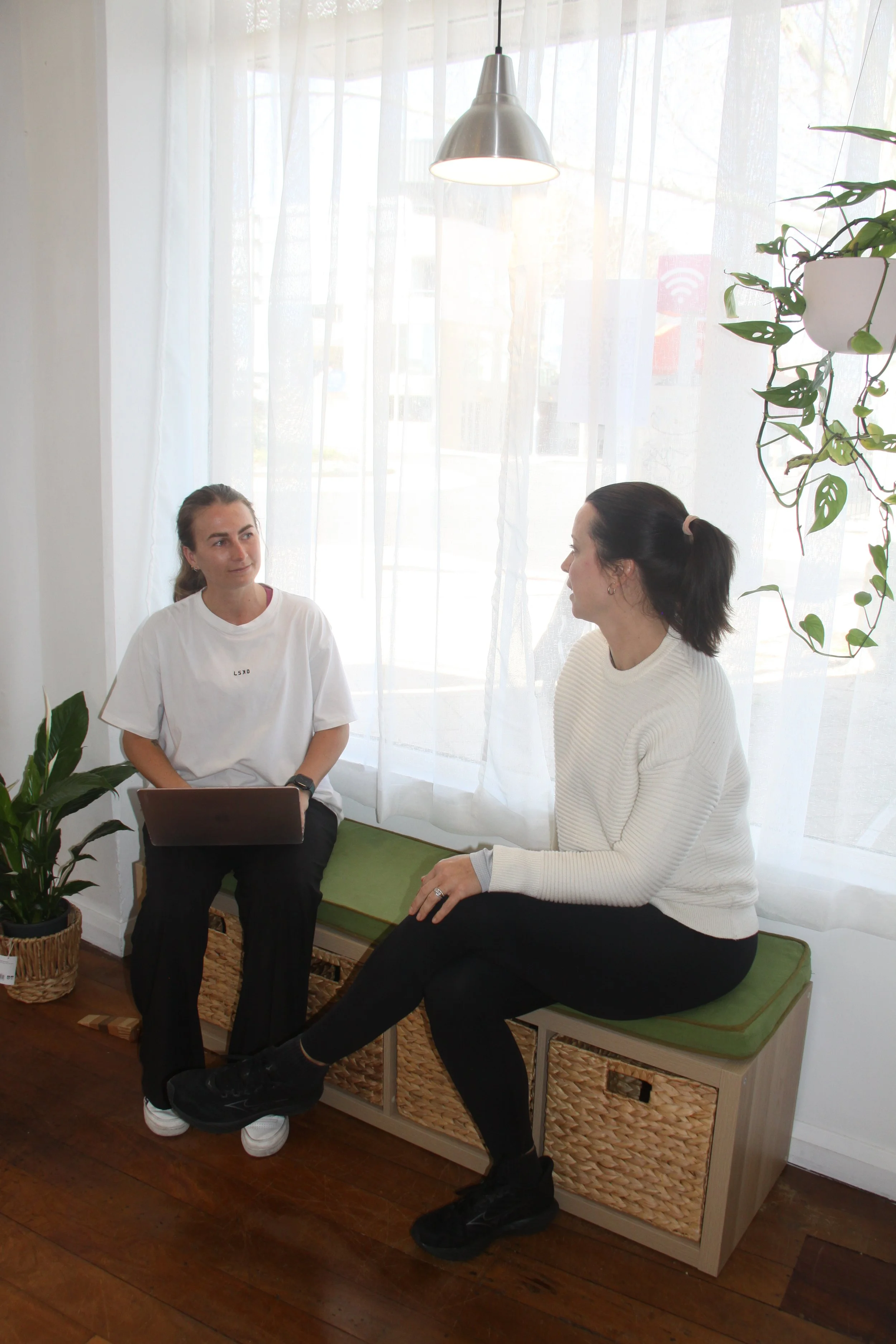 Two women engaging in a conversation in a bright room with sheer curtains, wooden flooring, and indoor plants. One woman is seated on a small bench with a laptop on her lap, and the other is sitting on a cushioned bench, facing her.