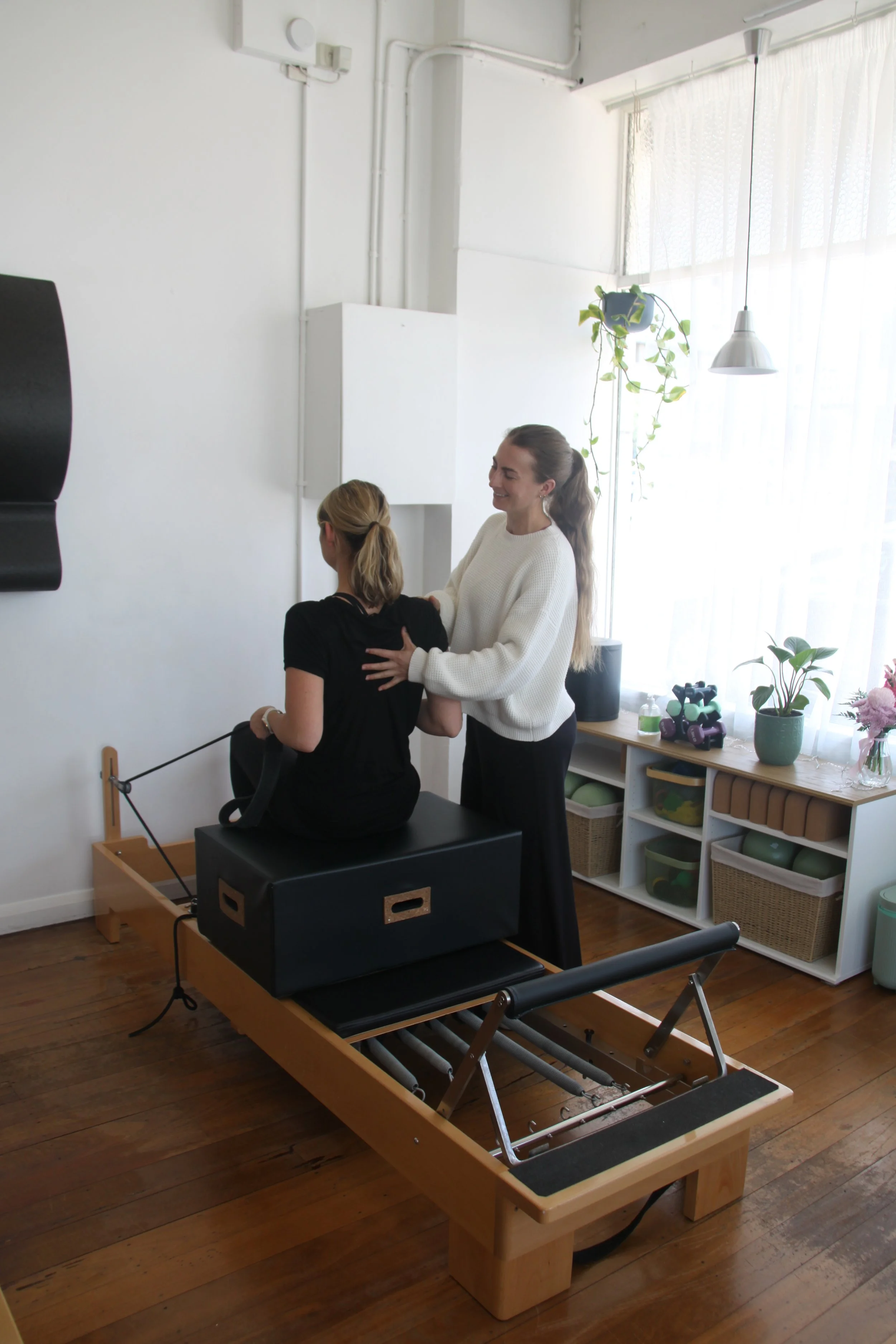 Two women are in a treatment room with a Pilates reformer machine. One woman is sitting on the chair attachment on the reformer, and the other woman is standing beside her, helping her. The room has white walls, wooden flooring, and natural sunlight coming through large windows with curtains. There are plants and gym equipment on a shelf in the background.