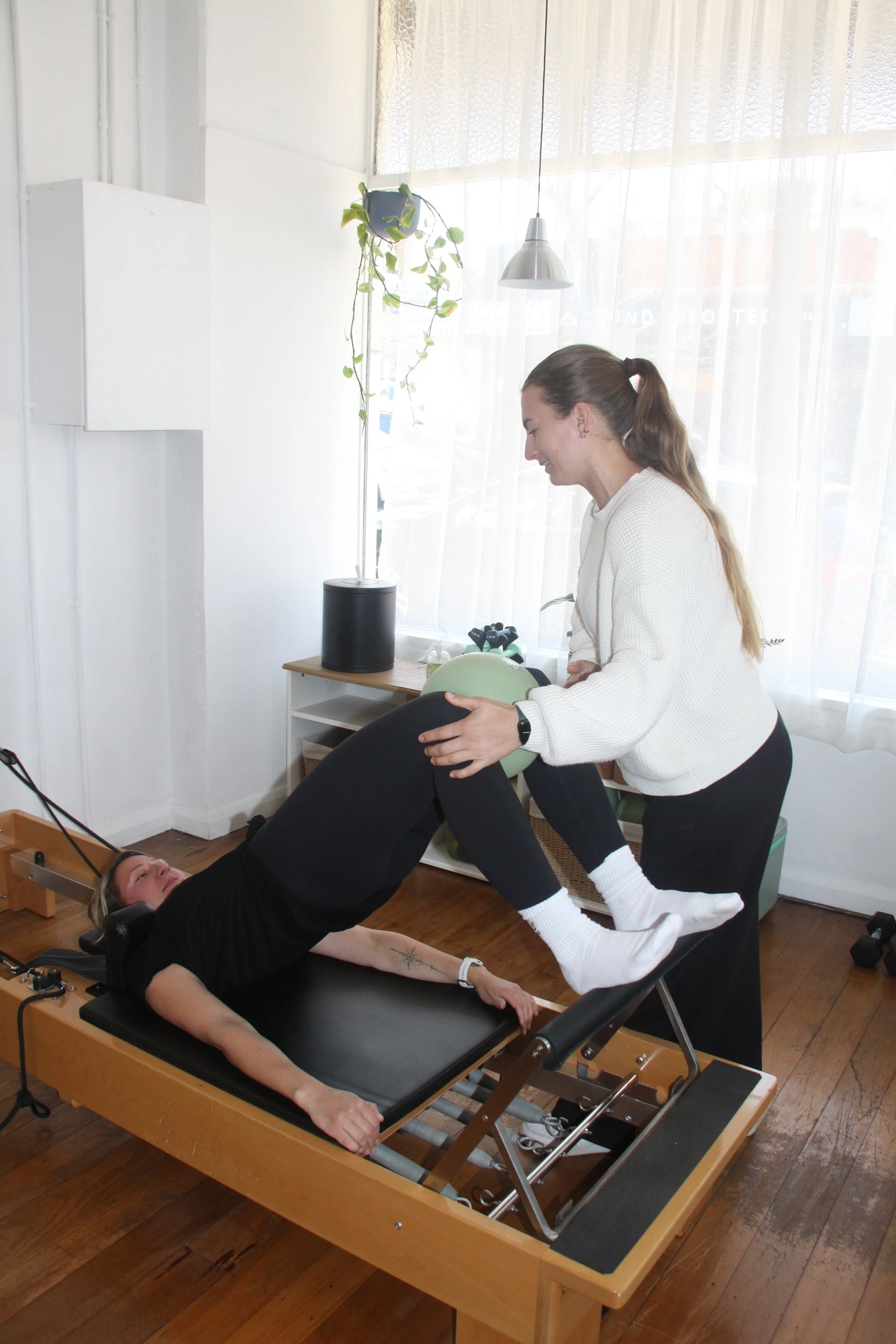 A woman lying on a chiropractic adjustment table with her legs raised by a practitioner. The practitioner is holding her legs and appears to be performing an exercise or chiropractic adjustment. The room has large windows with sheer curtains, a potted plant, and a hanging light.