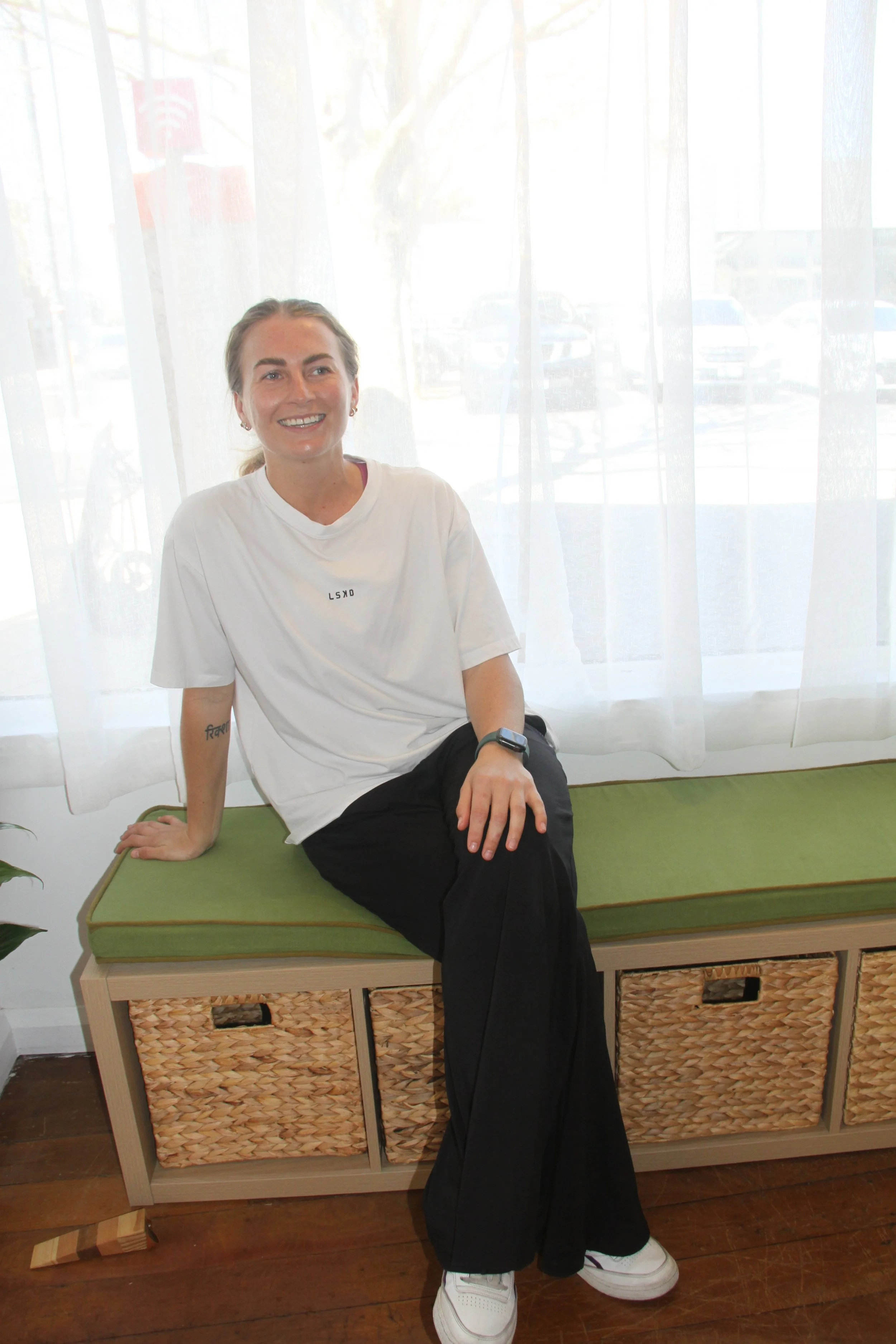 A young woman with blonde hair tied back, wearing a white t-shirt and black pants, sitting on a green cushion on a wooden bench with woven baskets in the drawers underneath, smiling and looking to her left. She is indoors near a window with sheer white curtains, and parked cars are visible outside.