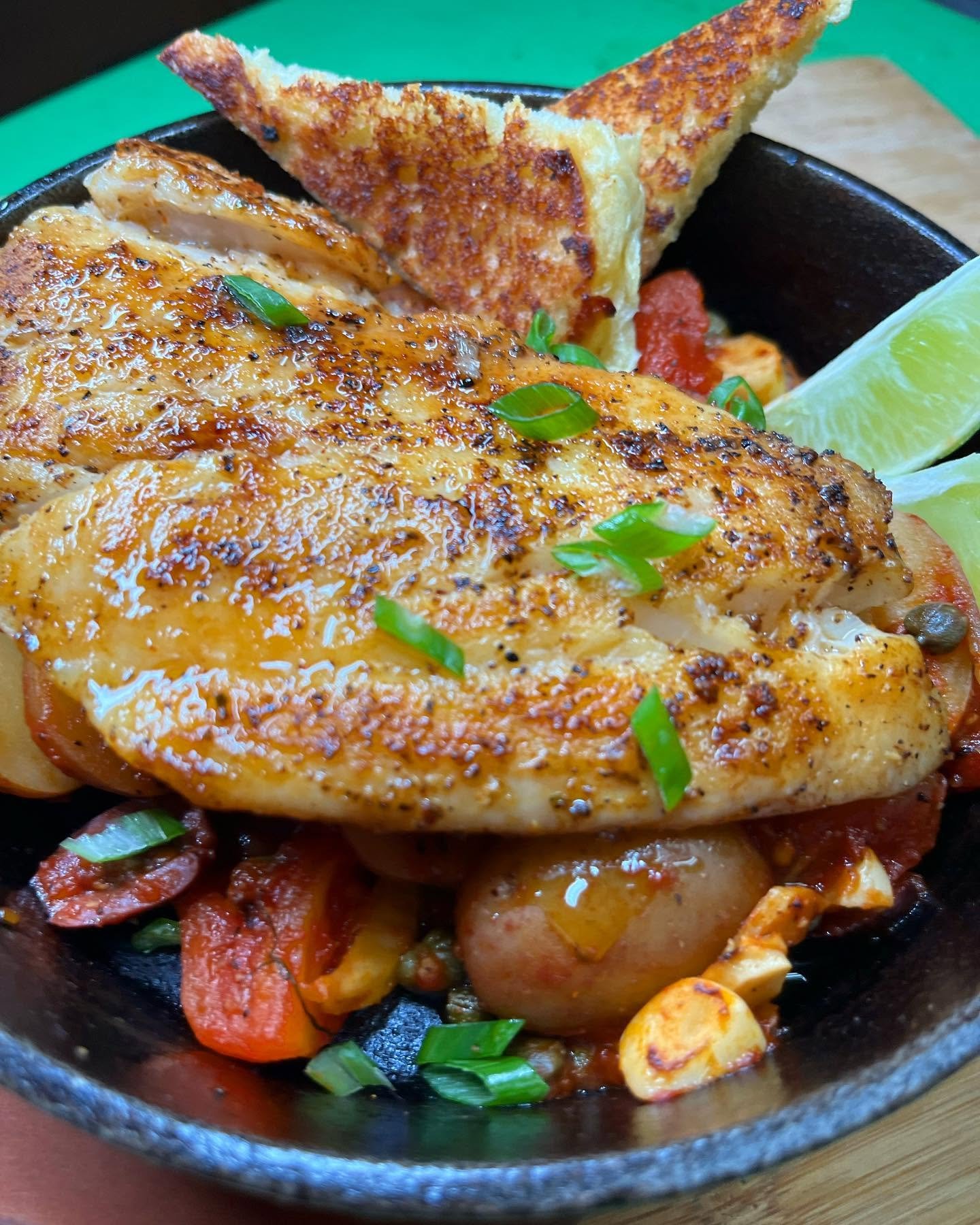Close-up of baked chicken breast topped with cheese and green onions, served with roasted potatoes, sliced tomatoes, a lemon wedge, and bread in a black skillet.