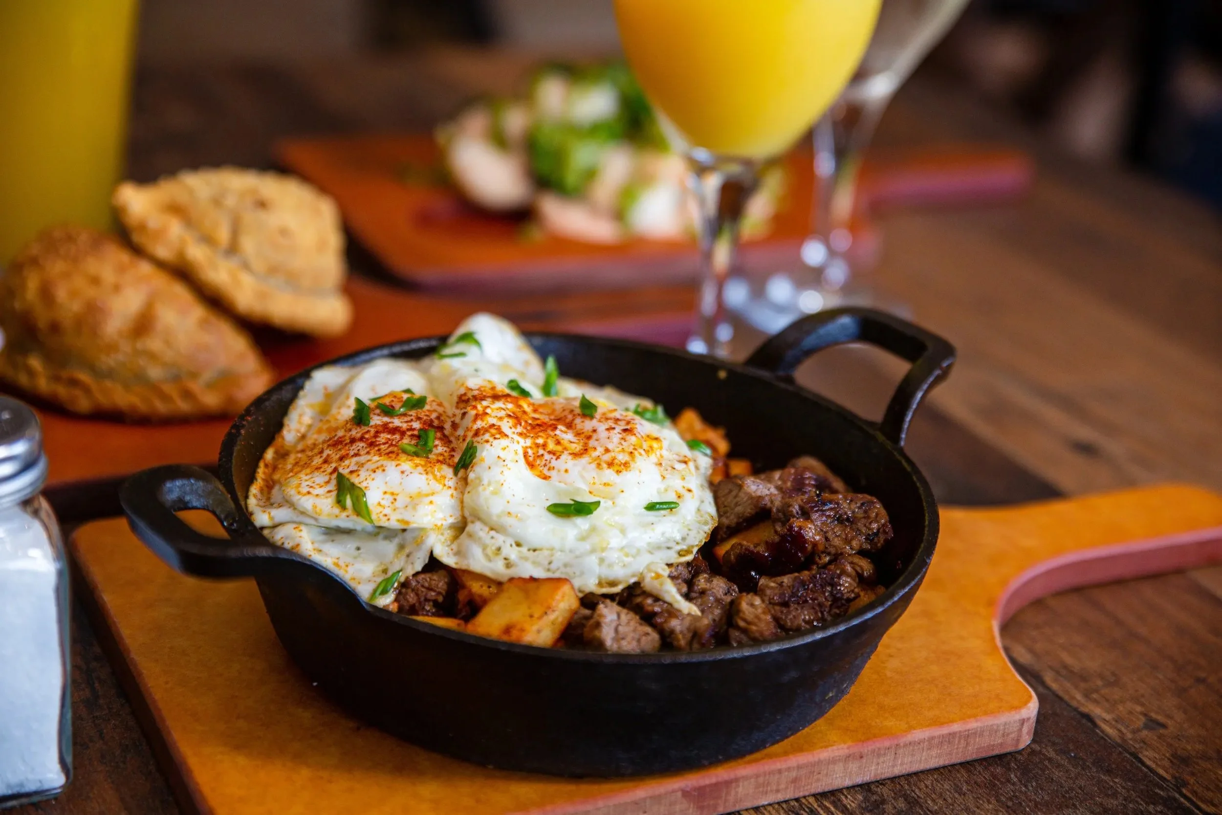 A sizzling skillet of breakfast dish with fried eggs topped with green herbs, served with cooked beef, and located on a wooden tray. In the background, there is a glass of yellow juice, pastries, a salad, and salt and pepper shakers on a dark wood ta