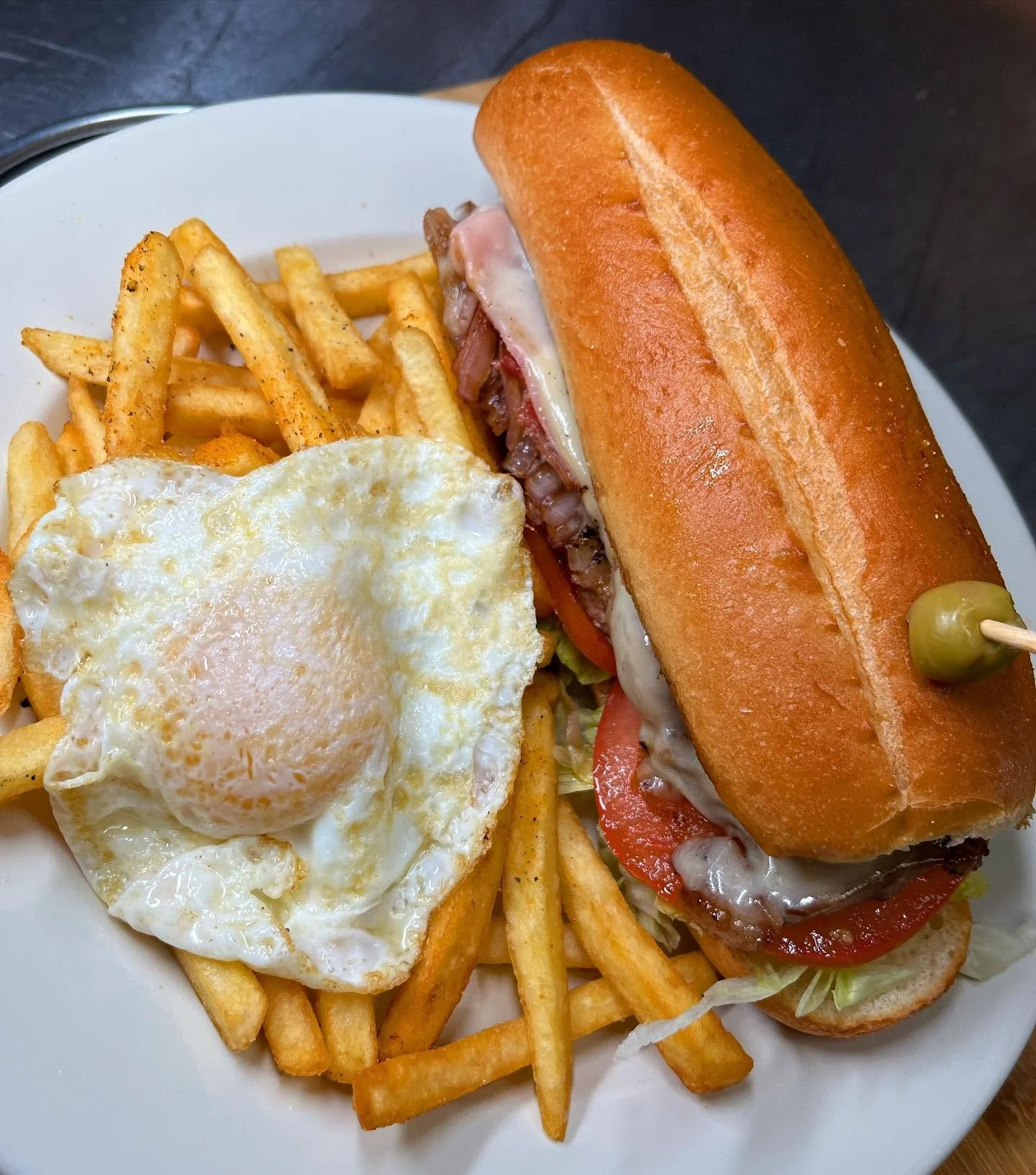 Plate with French fries, fried egg, and sandwich in a bun with sliced tomato, bacon, and cheese, garnished with a pickle