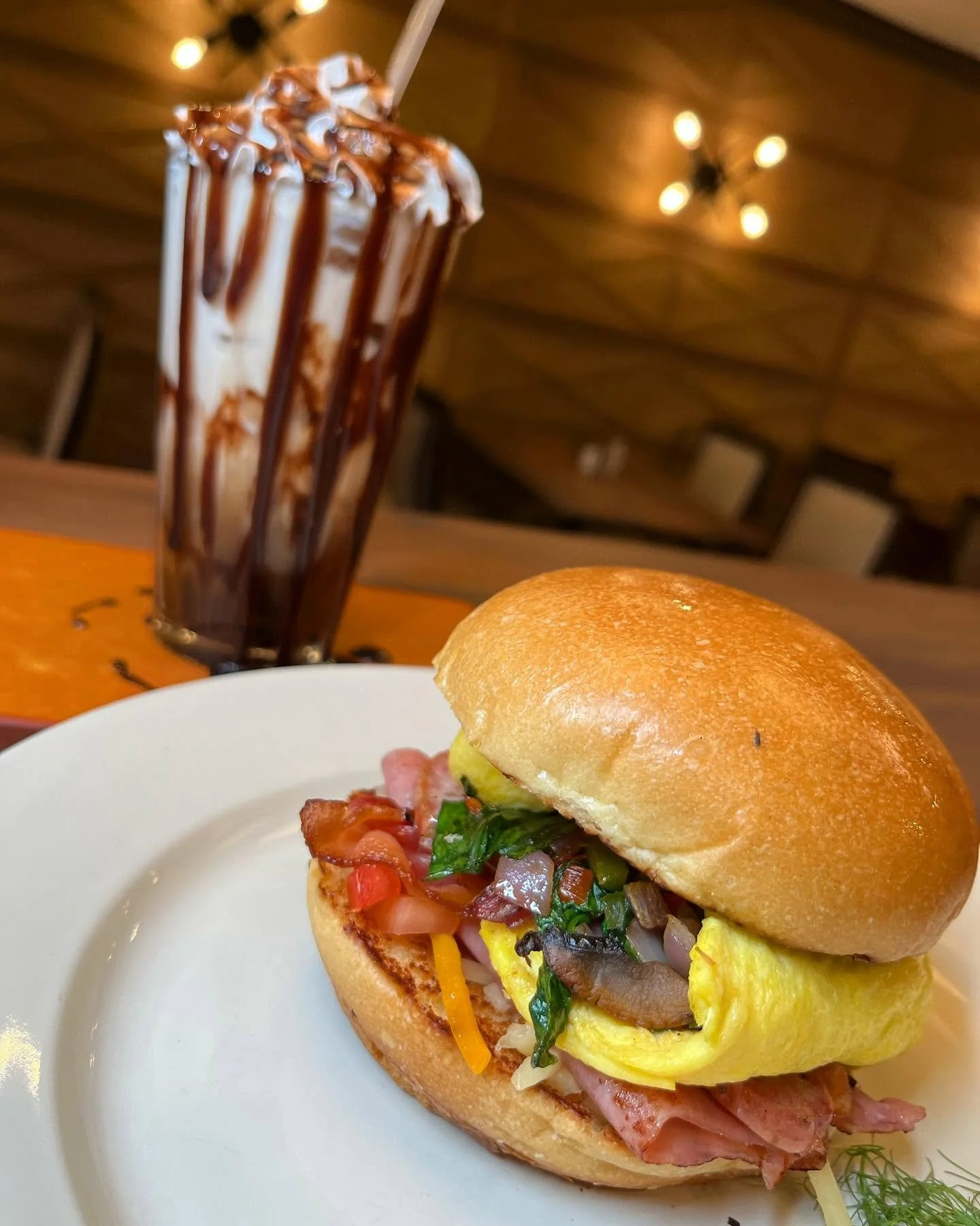 A plate with a sandwich featuring bacon, eggs, cheese, and vegetables, with a milkshake topped with chocolate syrup in the background.