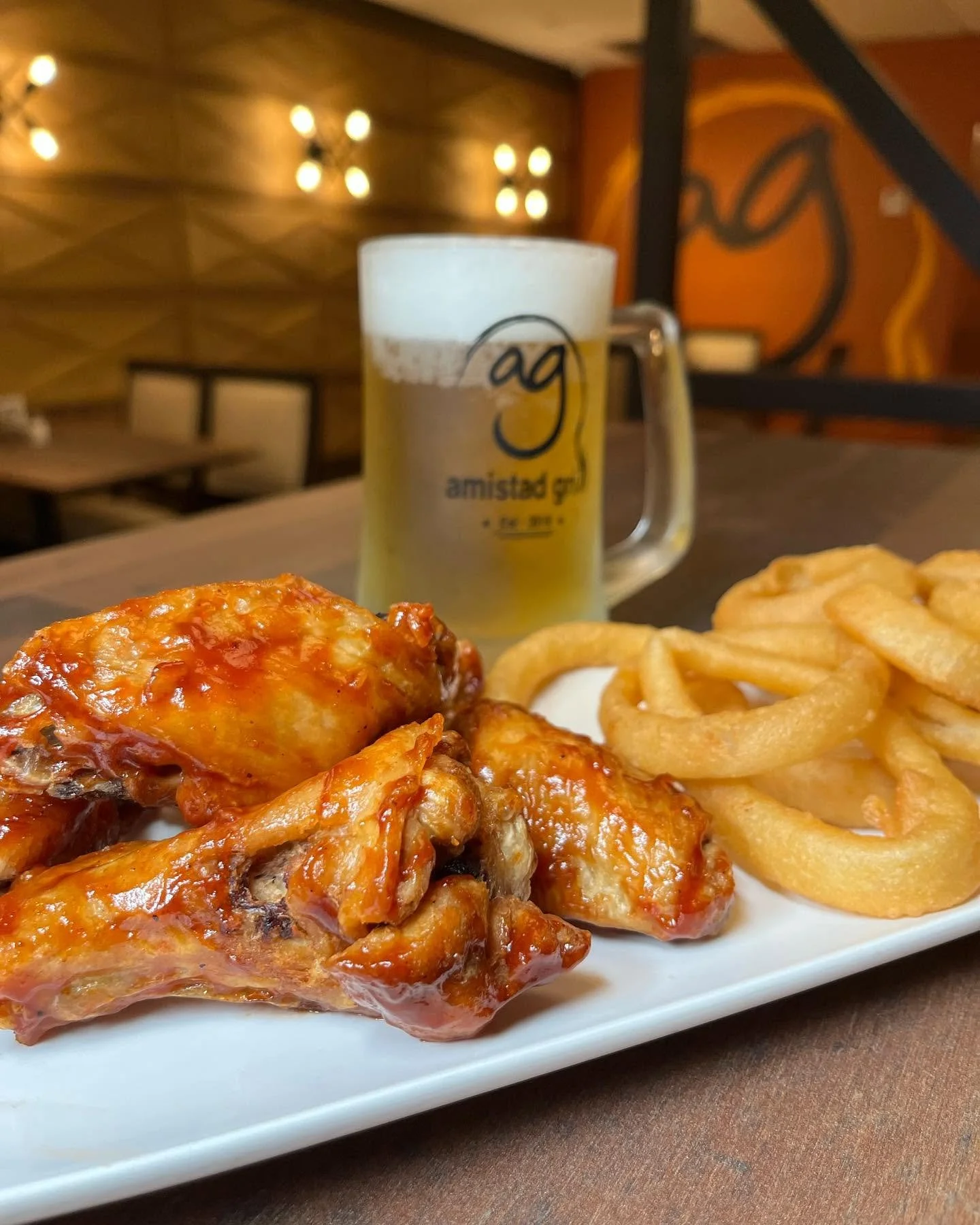 Plate with chicken wings covered in spicy orange sauce and a side of curly fries in front of a beer mug with beer, in a restaurant setting.