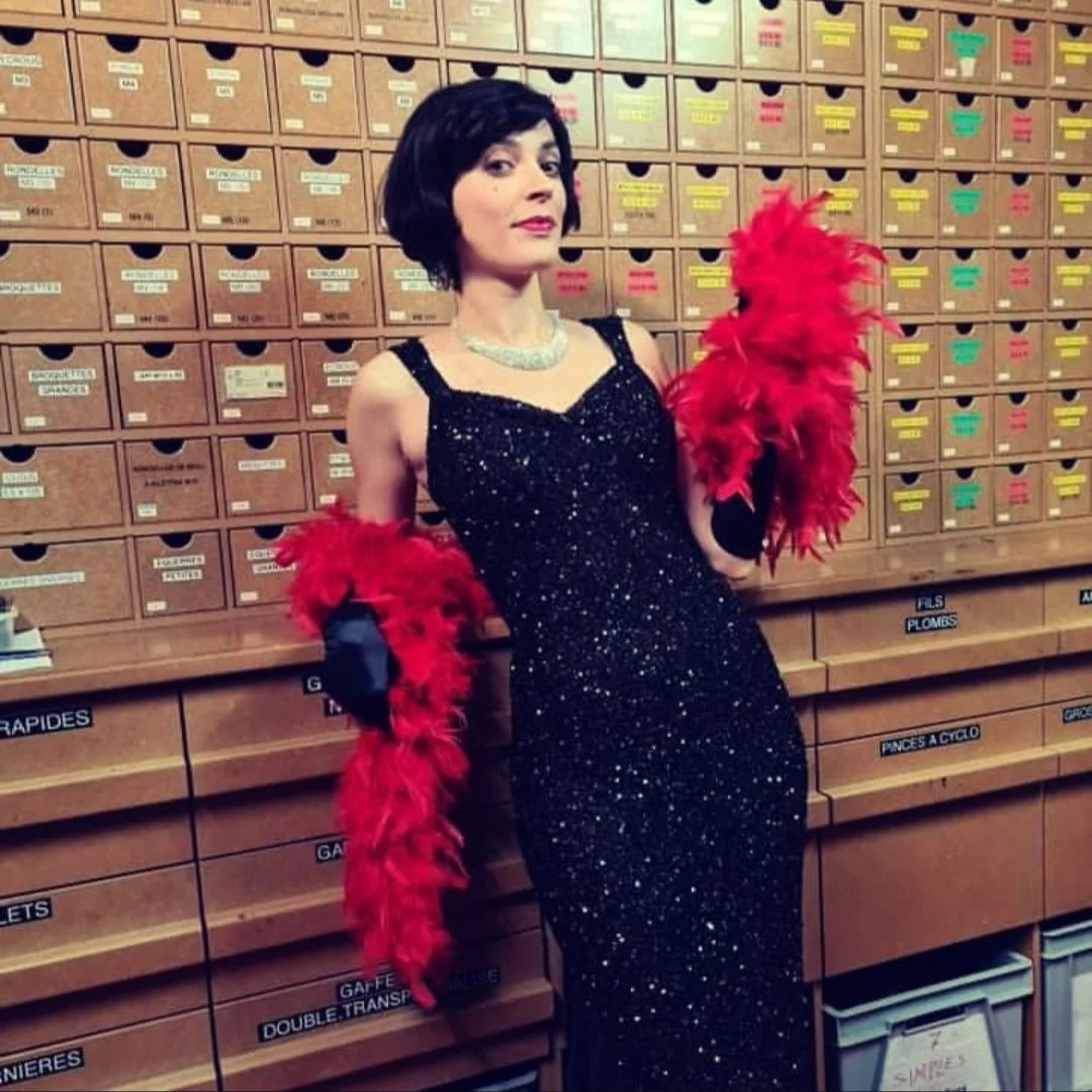 A woman in a black sparkly dress and pearl necklace posing in front of a wooden mail sorting cabinet, holding red feathered gloves.