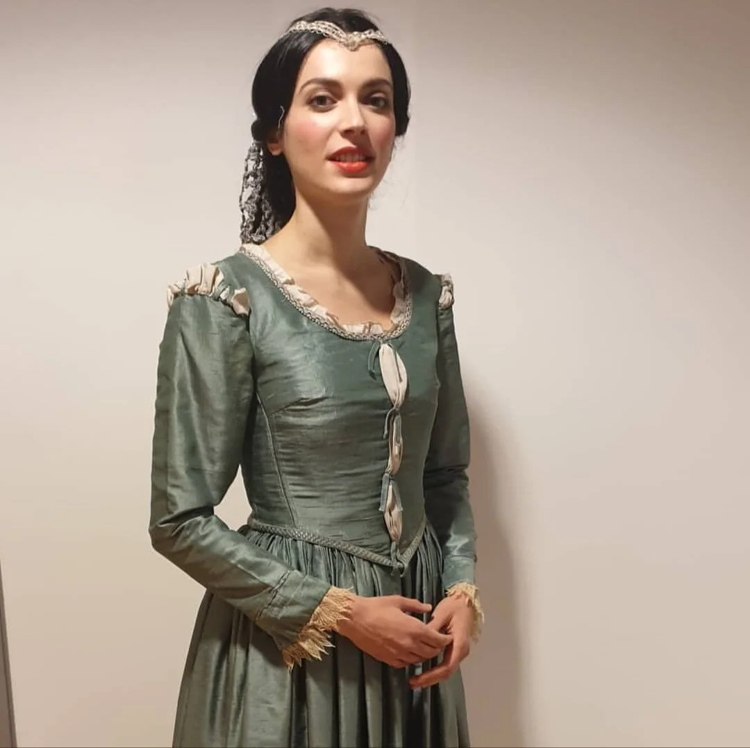 A woman dressed in a historical or medieval-style gown with long sleeves, lace accents, and a fitted waist, standing against a plain wall.