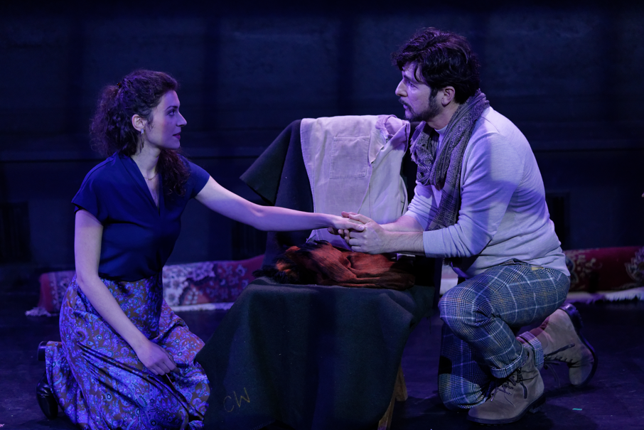A woman and a man are kneeling on the floor, holding hands and looking at each other, in a dimly lit room with a dark background.