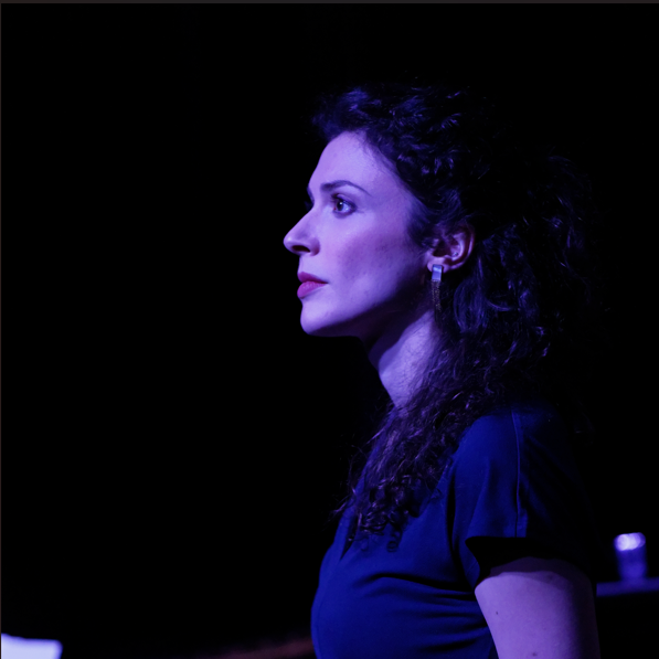 Profile of a woman with curly dark hair wearing a blue shirt and hoop earrings against a dark background.