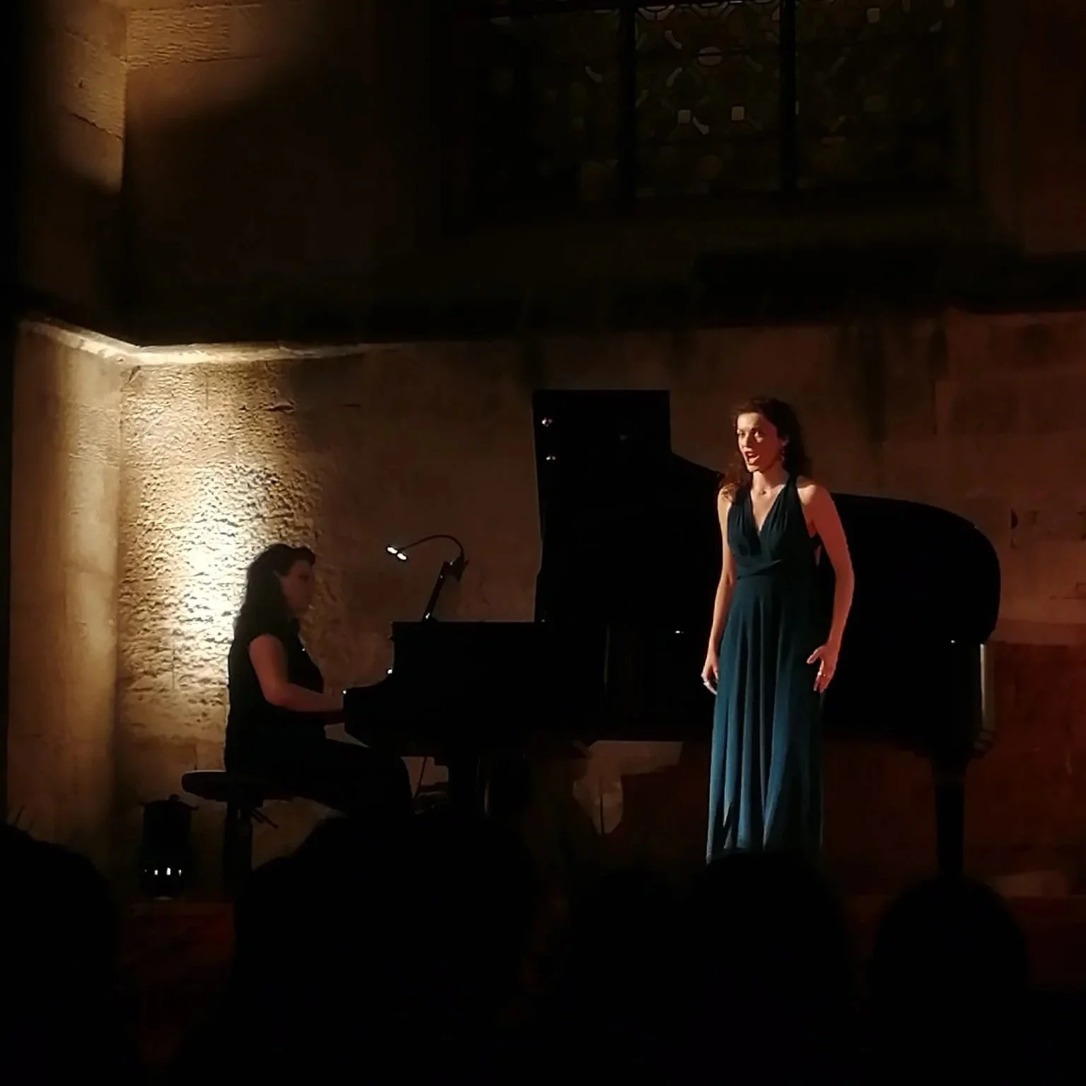 A woman in a long, blue dress singing at a concert, standing next to a grand piano with a pianist playing on it in a dimly lit room.