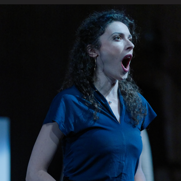 A woman with curly dark hair wearing a blue blouse, with her mouth open as if shouting or singing, against a dark background.
