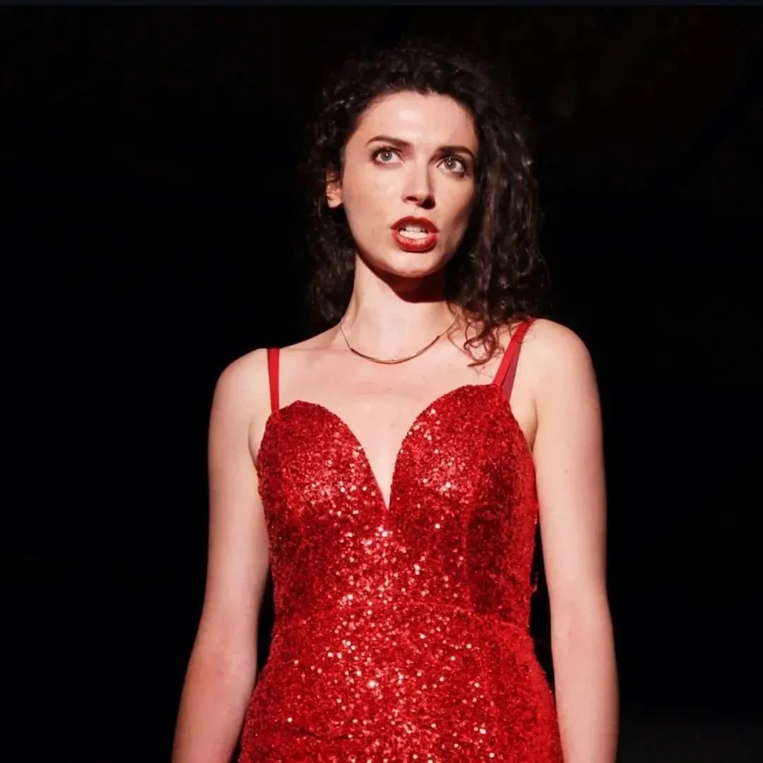 A woman with curly dark hair wears a red, sparkly, sleeveless dress with thin straps and a heart-shaped neckline, standing against a black background.