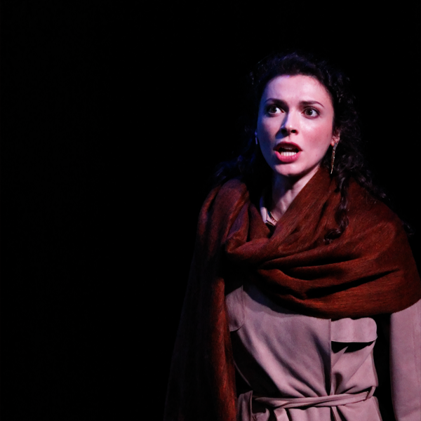 A woman with dark, curly hair and earrings is wearing a brown shawl and a beige dress. She appears to be on stage, with a dark background, and has an intense or emotional expression.