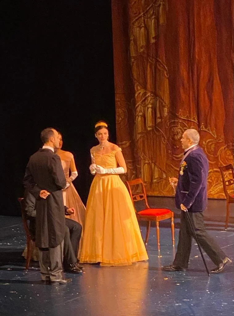 Stage scene with four actors in period costumes, two women in elegant gowns and two men in formal attire, one with a cane, on a theater stage with a decorative backdrop.