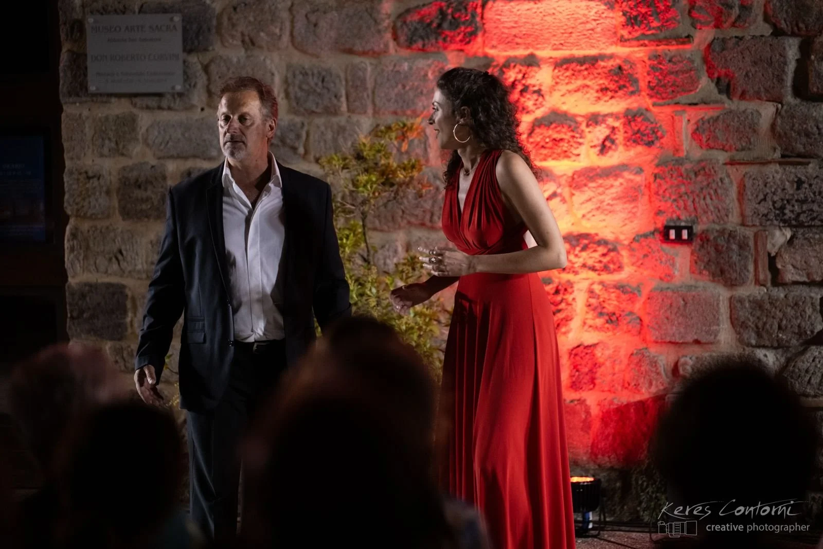 Two people, a man in a black suit and a woman in a red dress, standing and talking in front of a rustic brick wall with red lighting. Audience members are partially visible at the bottom.
