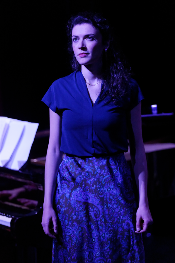 A woman with long, curly hair standing in front of a piano, wearing a blue blouse and patterned skirt, illuminated by stage lighting.