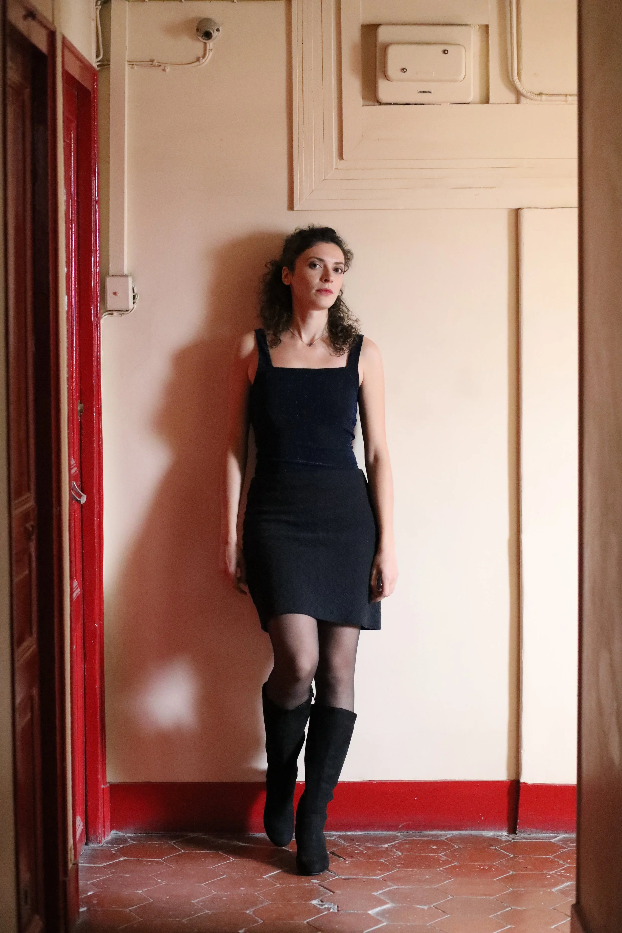 A woman with curly hair wearing a black dress, black stockings, and black boots standing against a beige wall with wooden panels, red trim, and an overhead electrical box.