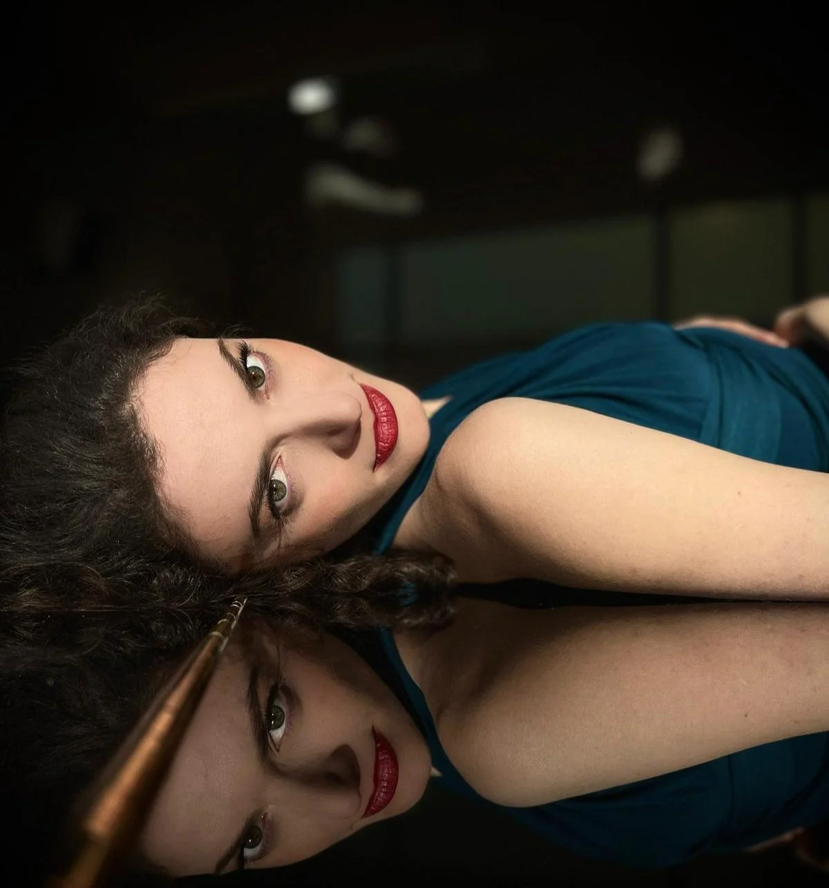 A woman with dark curly hair lying on her side in a dark room, wearing a teal dress, with reflection visible on a shiny surface beneath her, looking directly at the camera.