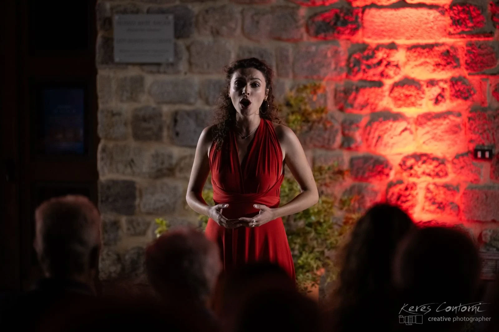 A woman in a red dress performing a song or speech on an indoor stage with a stone wall background and audience in the foreground.