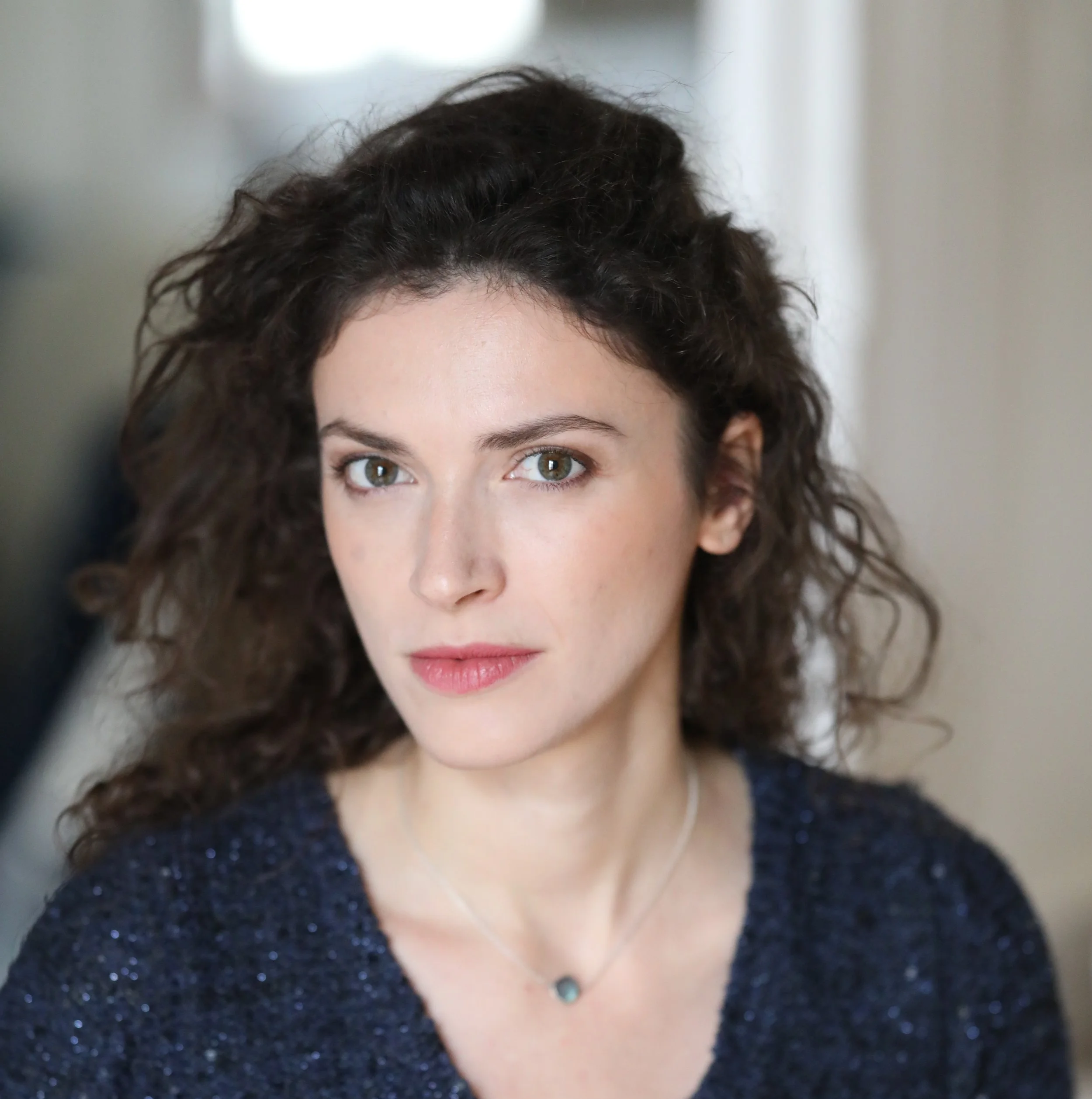 Close-up of a woman with curly dark hair and light skin, wearing a glittery black dress and a turquoise necklace, with her eyes looking directly into the camera.