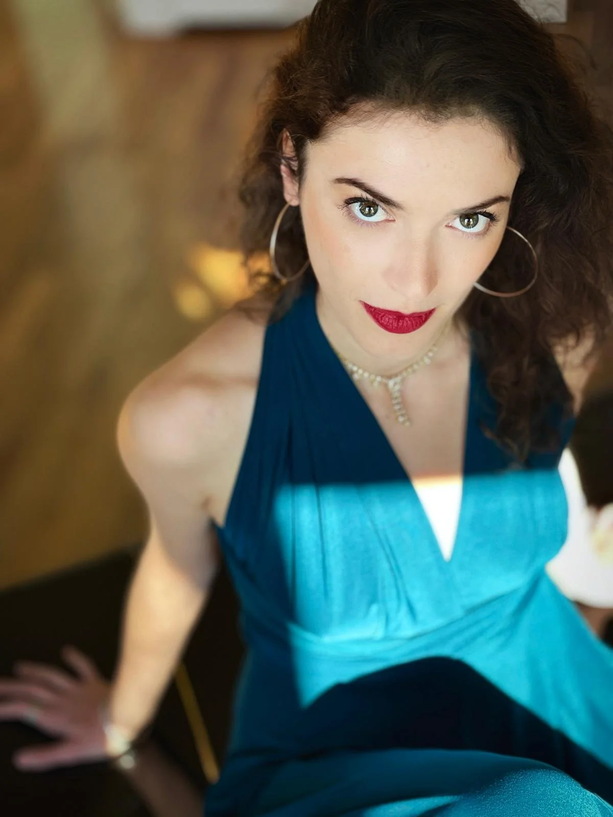 A woman with curly brown hair, blue eyes, and red lipstick, wearing a blue dress, hoop earrings, and a pearl necklace, looking up at the camera.