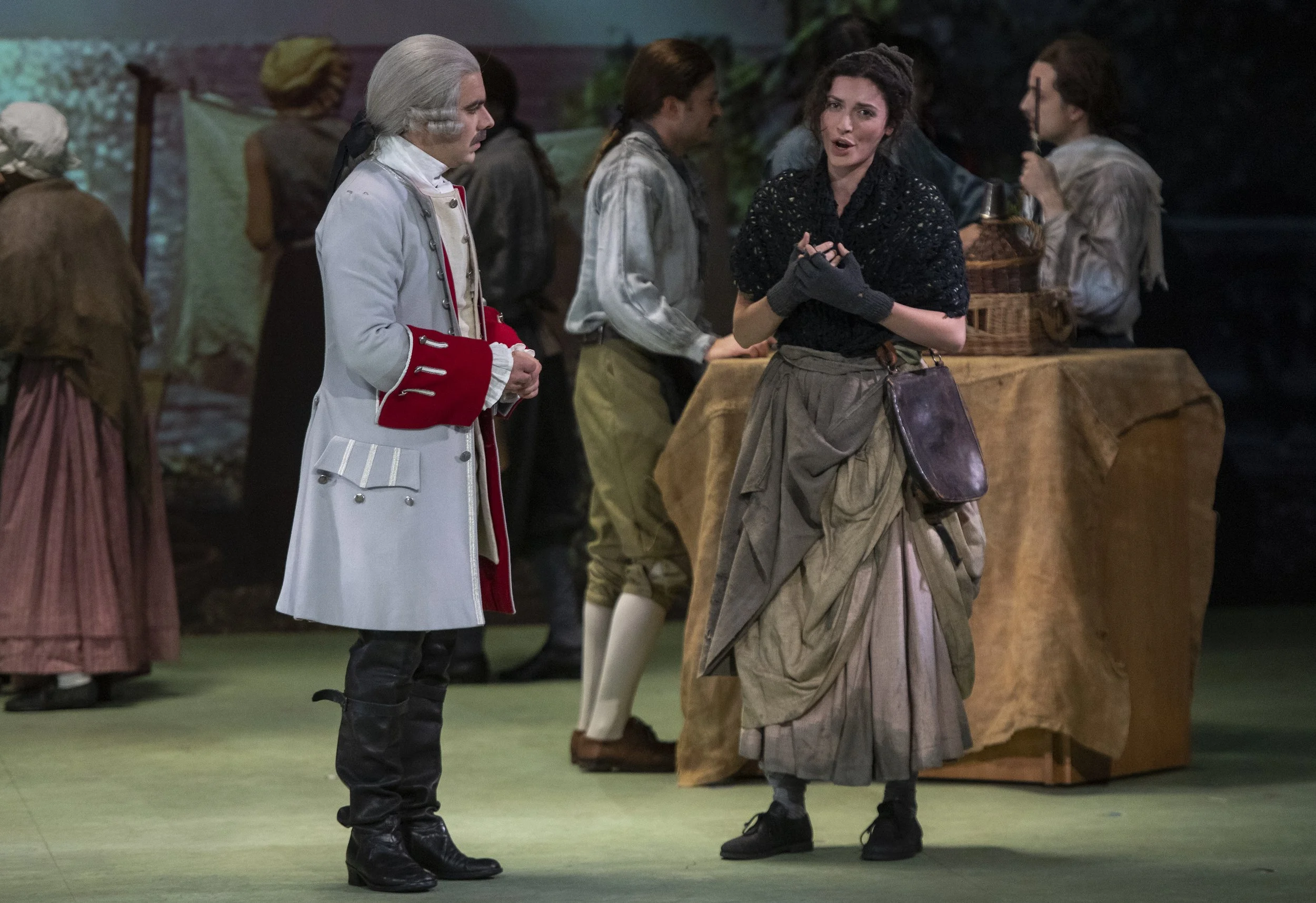 Actors in historical costumes on stage, with one woman talking and a man listening, during a theatrical performance.